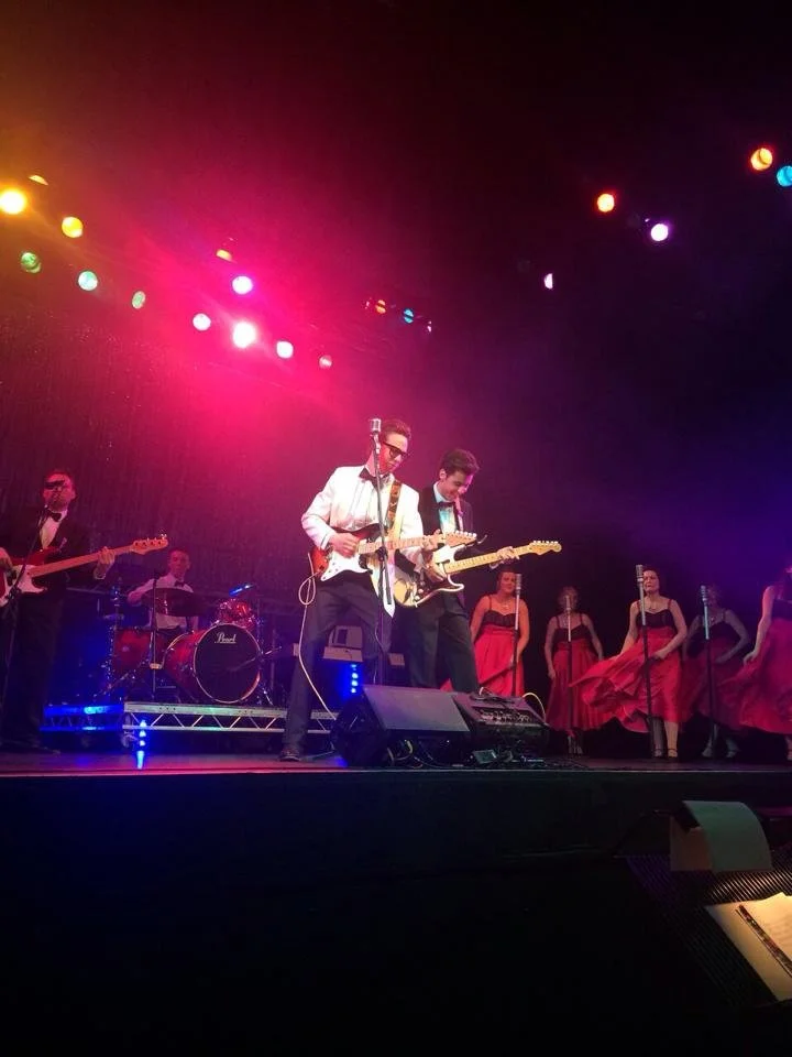 Musicians performing on stage with colorful stage lighting, including a guitarist singing into a microphone, a drummer in the background, other guitarists, and female backup singers in red dresses.