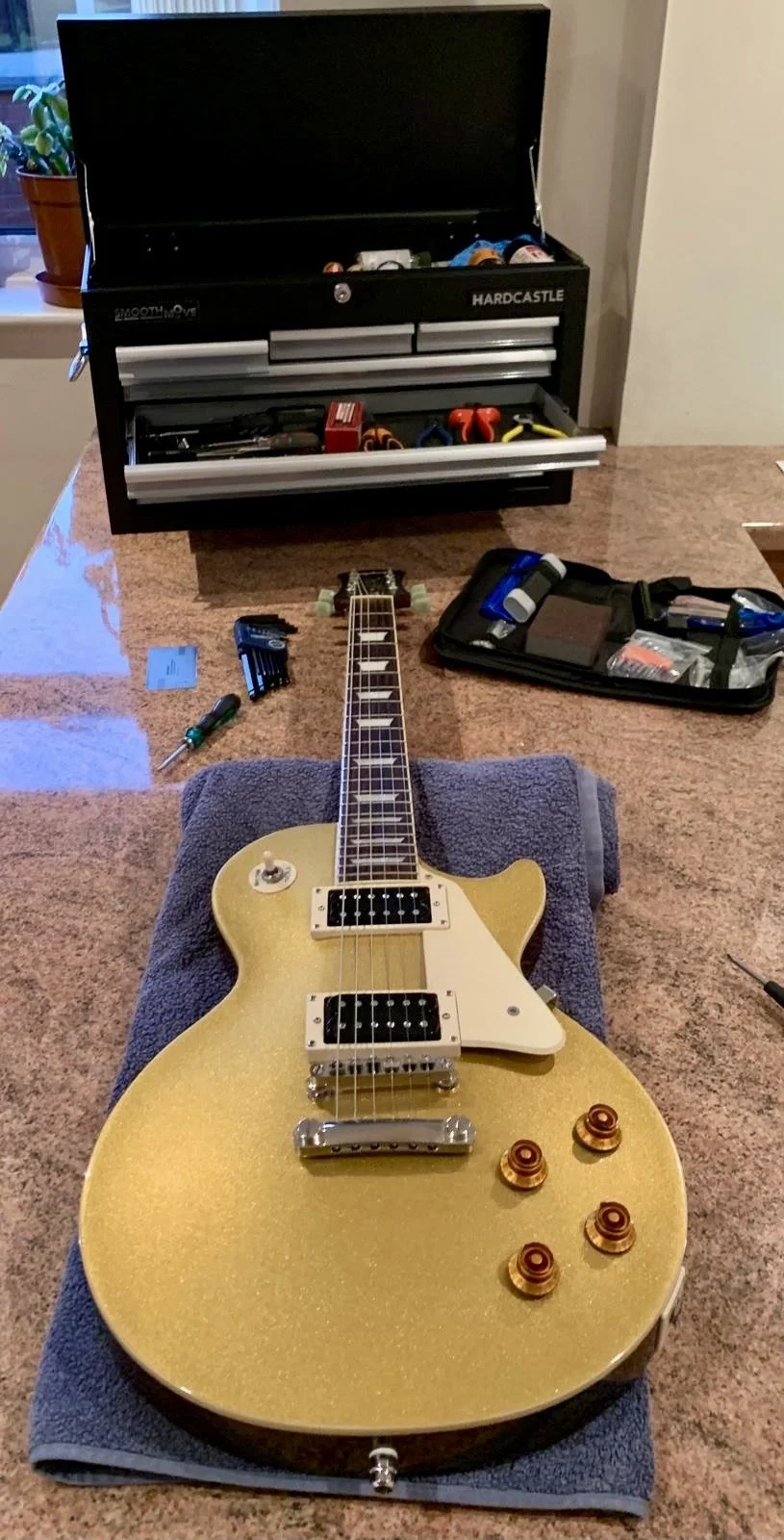 Gold electric guitar with four control knobs and open tuner on a towel, sitting on a table with a tool kit and open toolbox behind it.