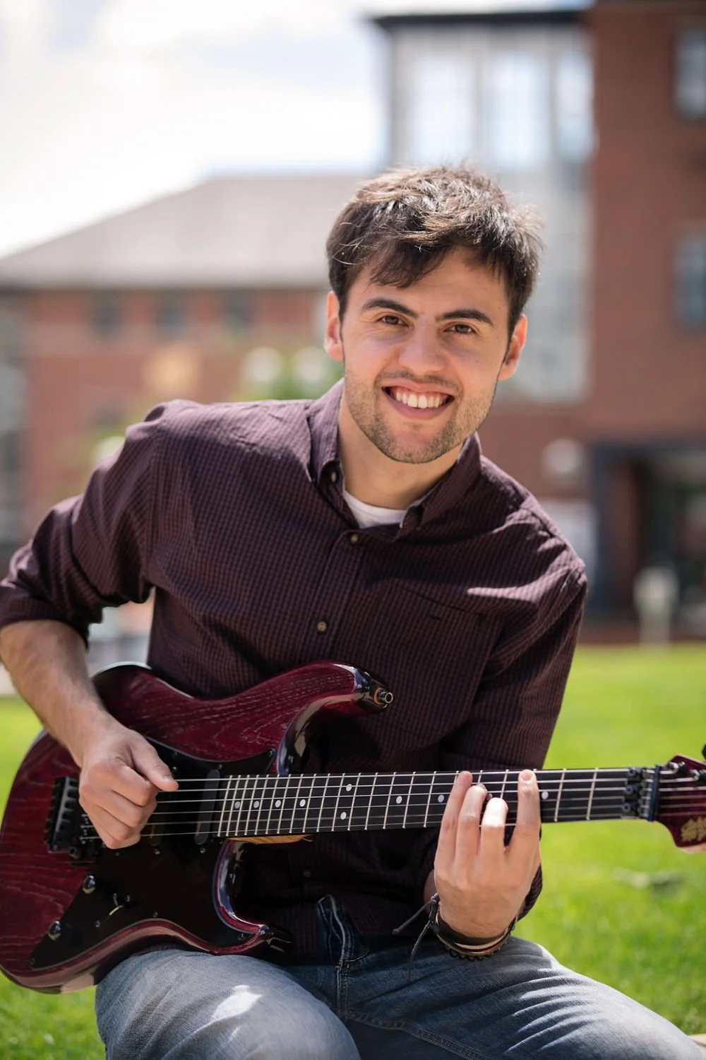 A young man with dark hair and a beard, smiling, and sitting outdoors on grass, playing an electric guitar.