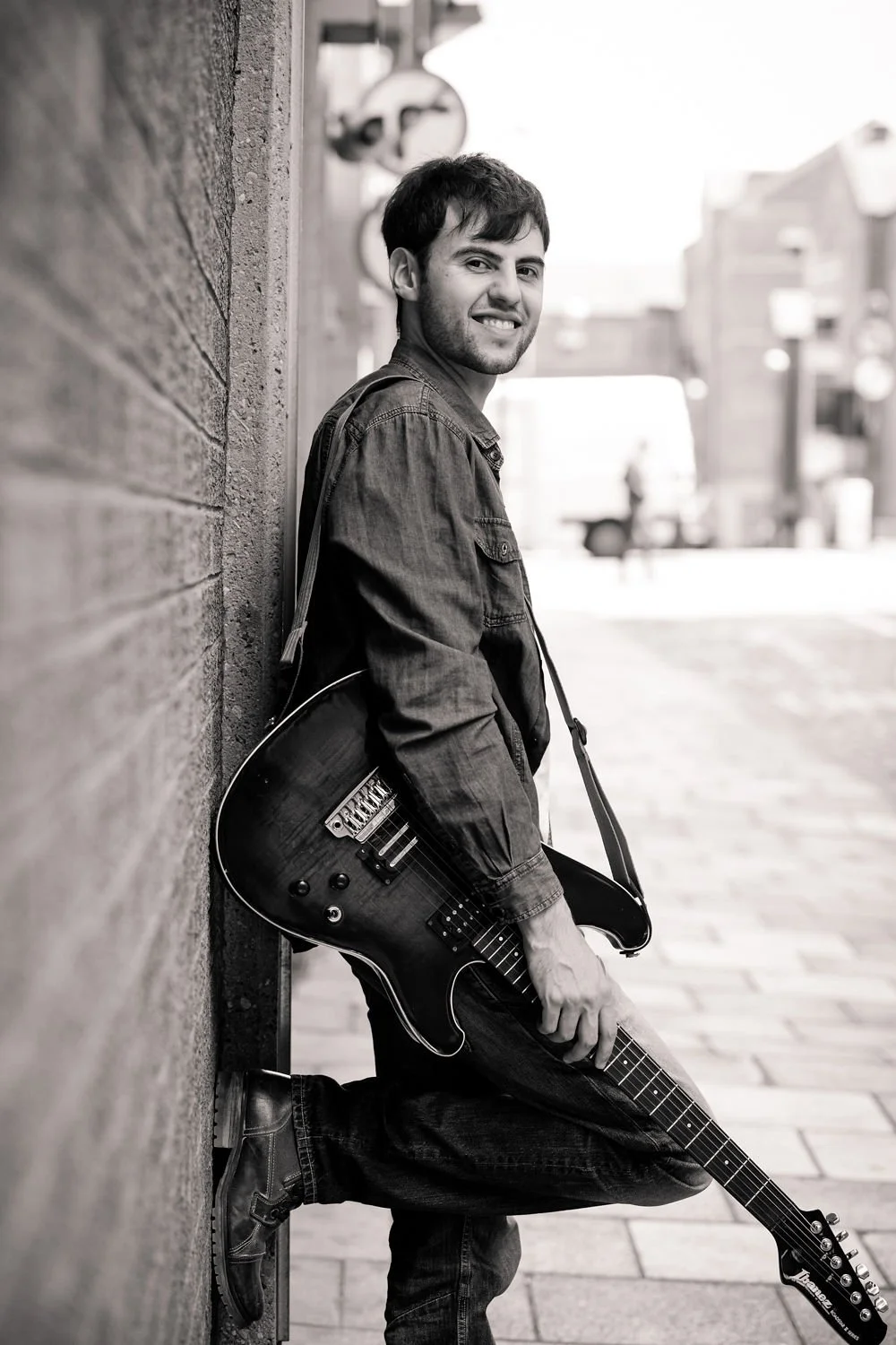A young man with dark hair and a beard, smiling while leaning against a brick wall on a city street, holding an electric guitar.