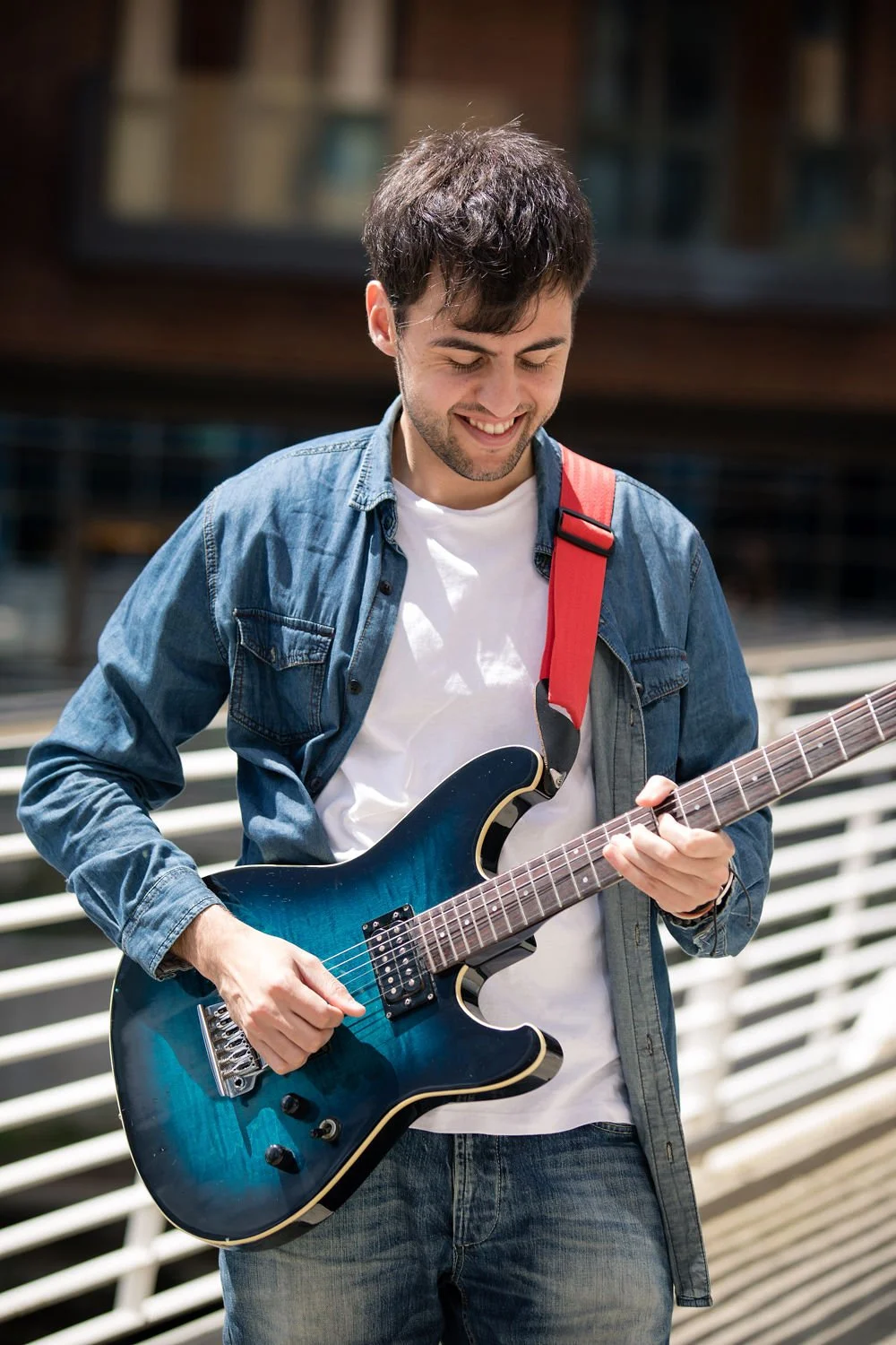 A young man with dark hair and a beard playing an electric guitar outdoors, smiling and wearing a denim jacket over a white t-shirt.