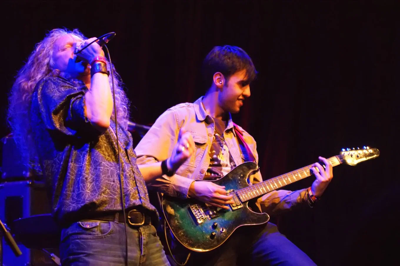 Two musicians performing on stage. The woman on the left is singing into a microphone and has curly blonde hair, wearing a patterned shirt. The man on the right is playing an electric guitar and has short dark hair, wearing a beige jacket.