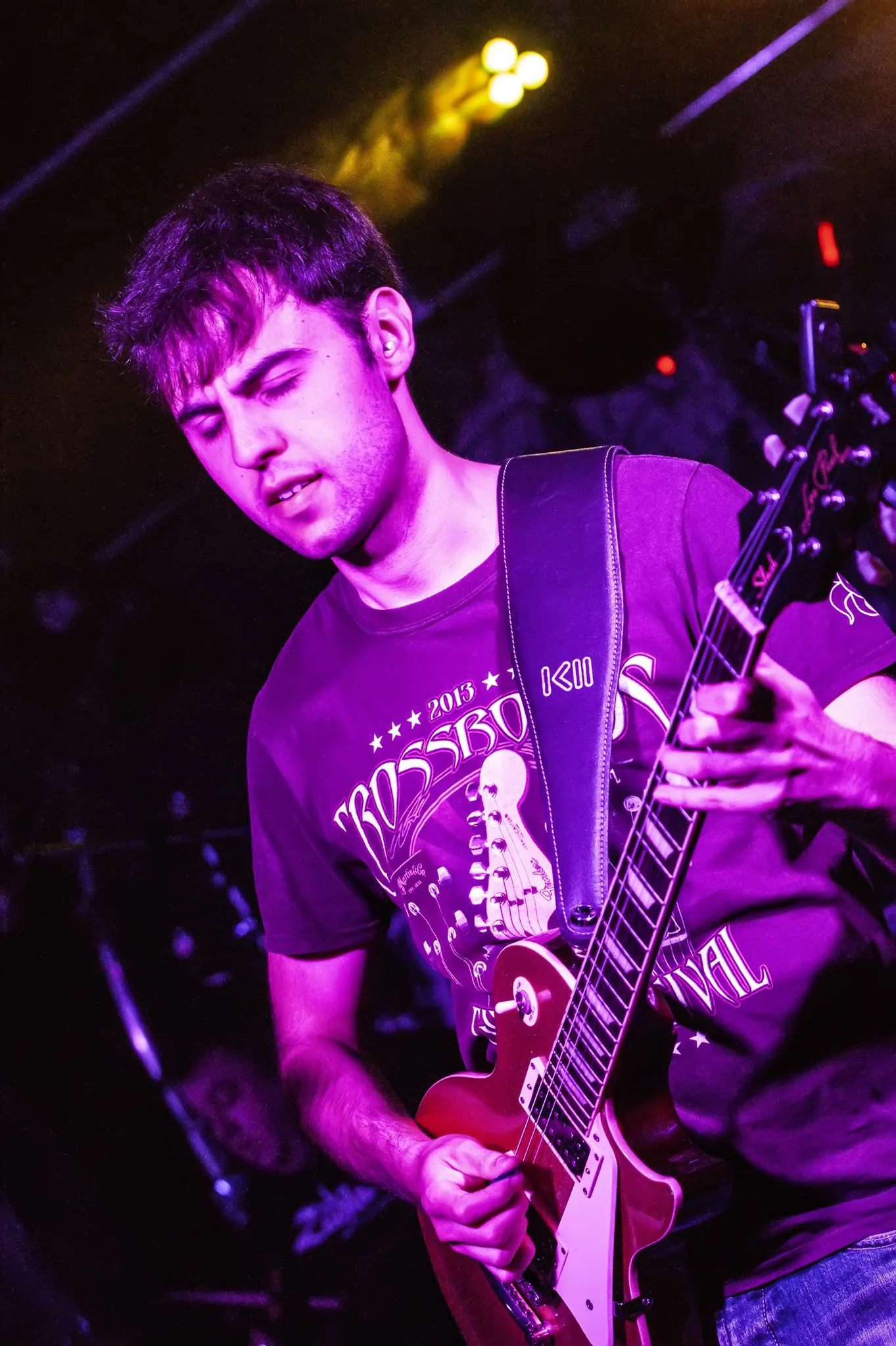 A young man playing an electric guitar on stage, illuminated by purple and yellow stage lights, with a focused or intense expression.