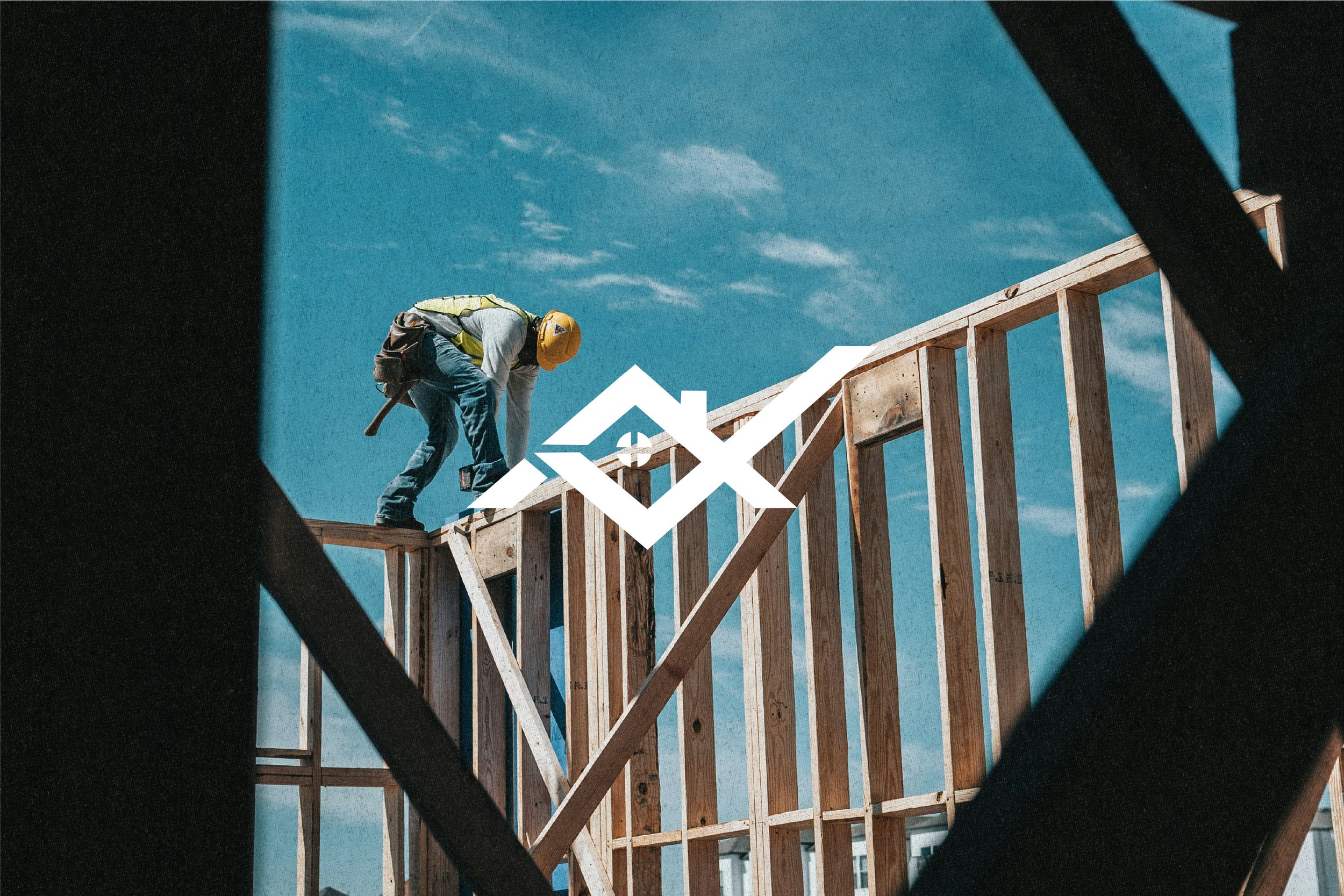 A construction worker wearing a yellow safety helmet and gray jacket working on the wooden frame of a building against a partly cloudy blue sky.