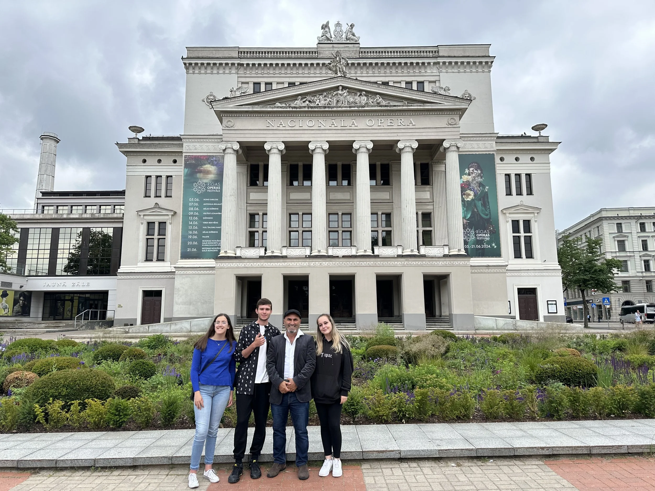 Four people standing in front of a large white neoclassical building with columns, labeled 'Nacionalā Opera', with banners on either side, a garden with bushes and flowers in the foreground, and cloudy sky overhead.