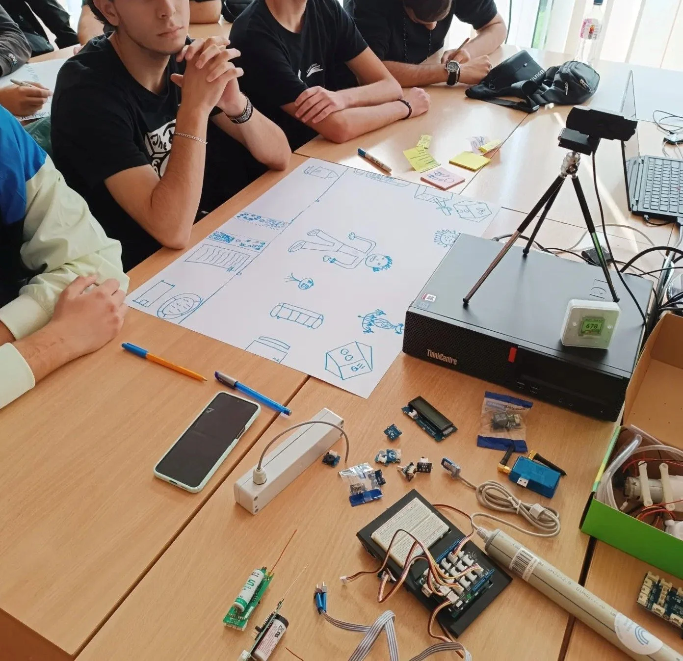 Group of students sitting around a table with a large drawing of a person and various objects on it, surrounded by electronics components, a smartphone, and a camera on a tripod.