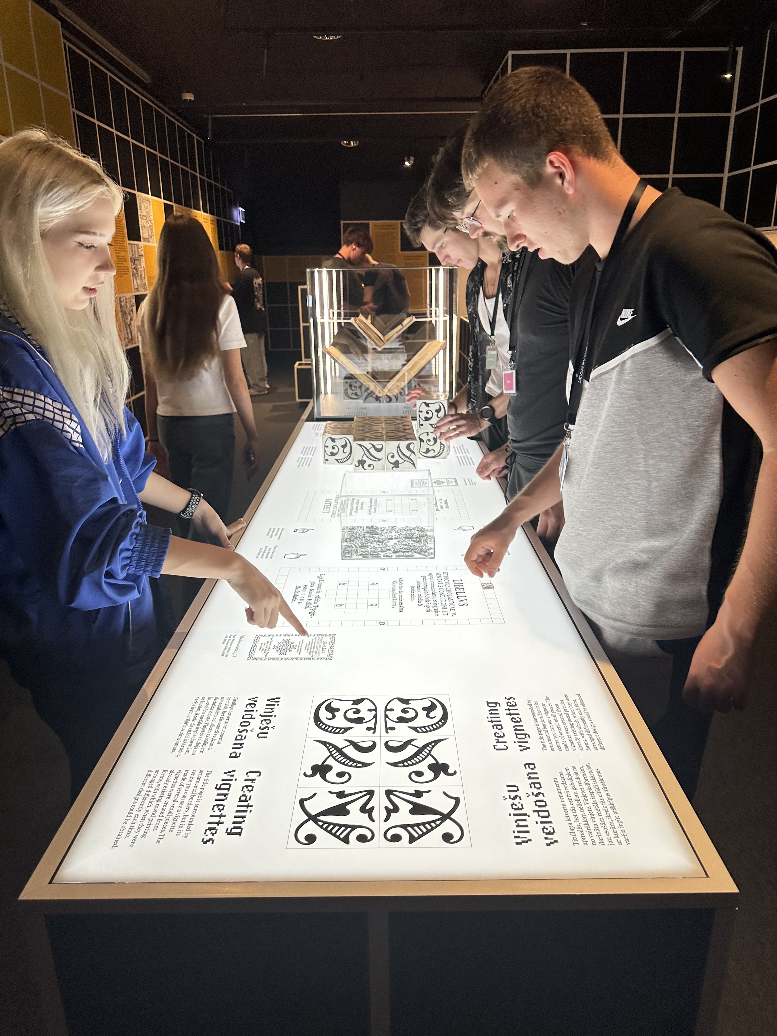 Group of people at a museum exhibit viewing an illuminated information display, with some pointing and reading.