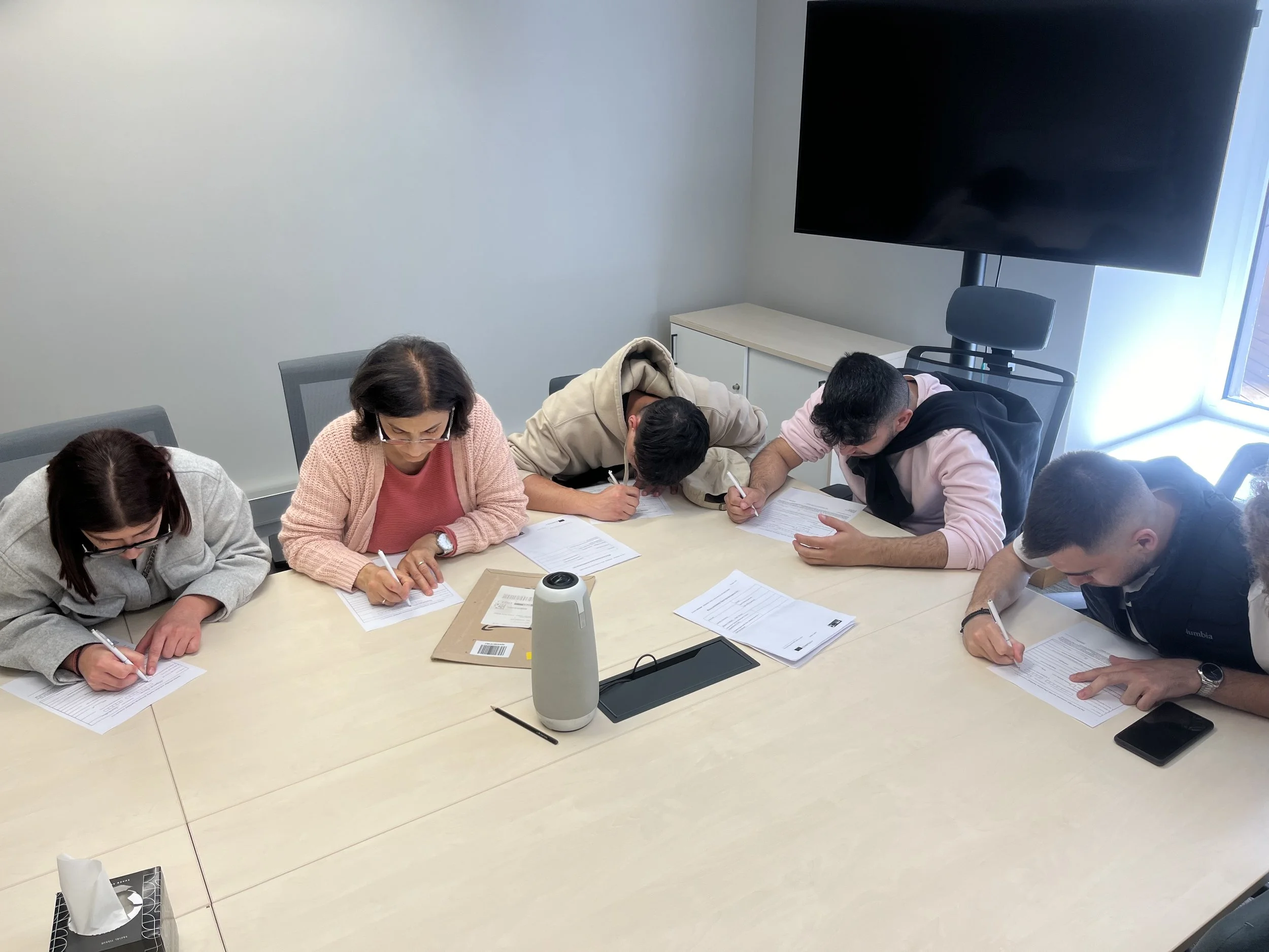 Six people sitting around a conference table, writing on papers in a meeting room with a large monitor on the wall.