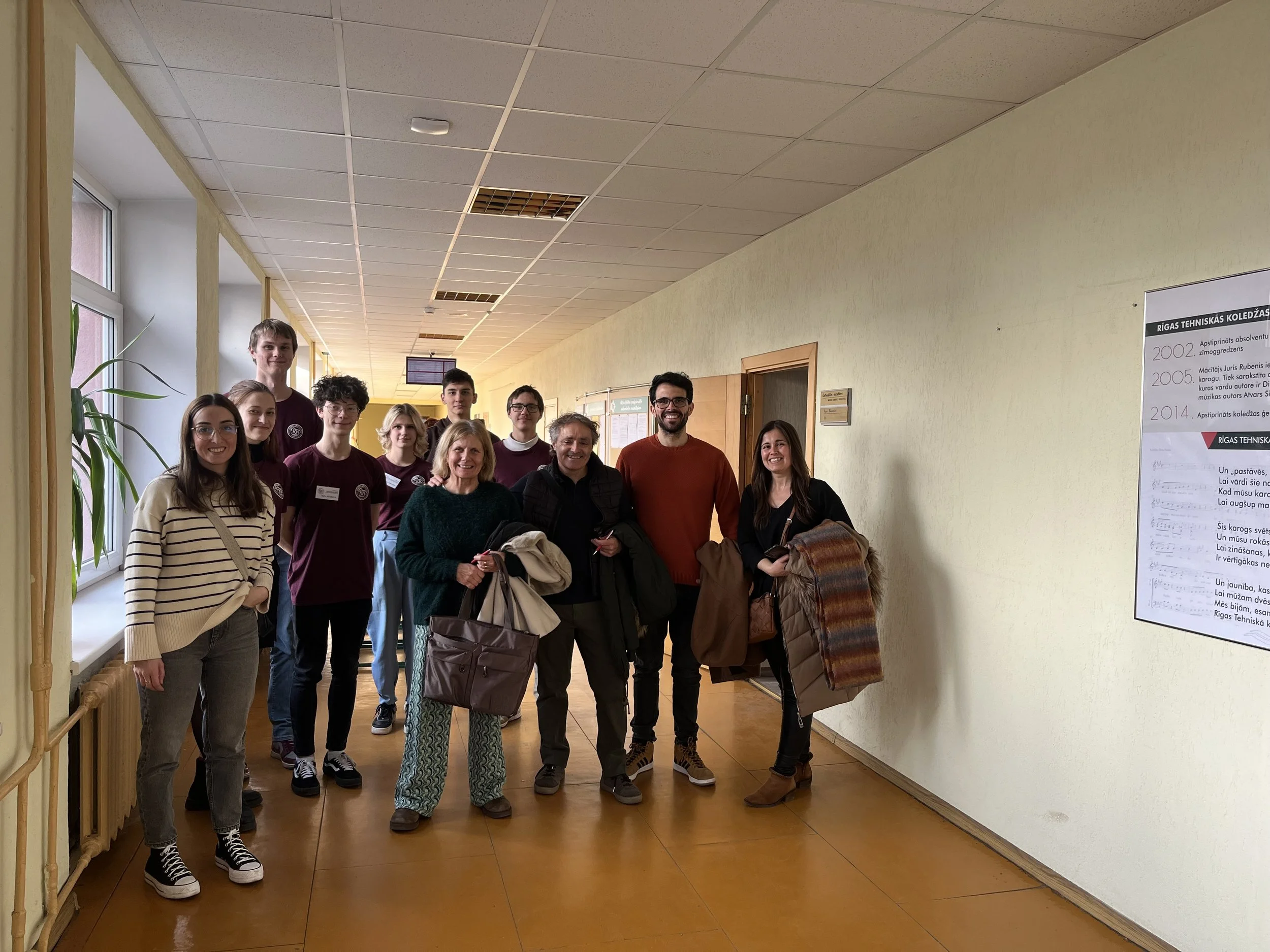A group of eleven people standing in a school corridor, smiling at the camera. There is a window with a plant on the left and a poster on the right wall.