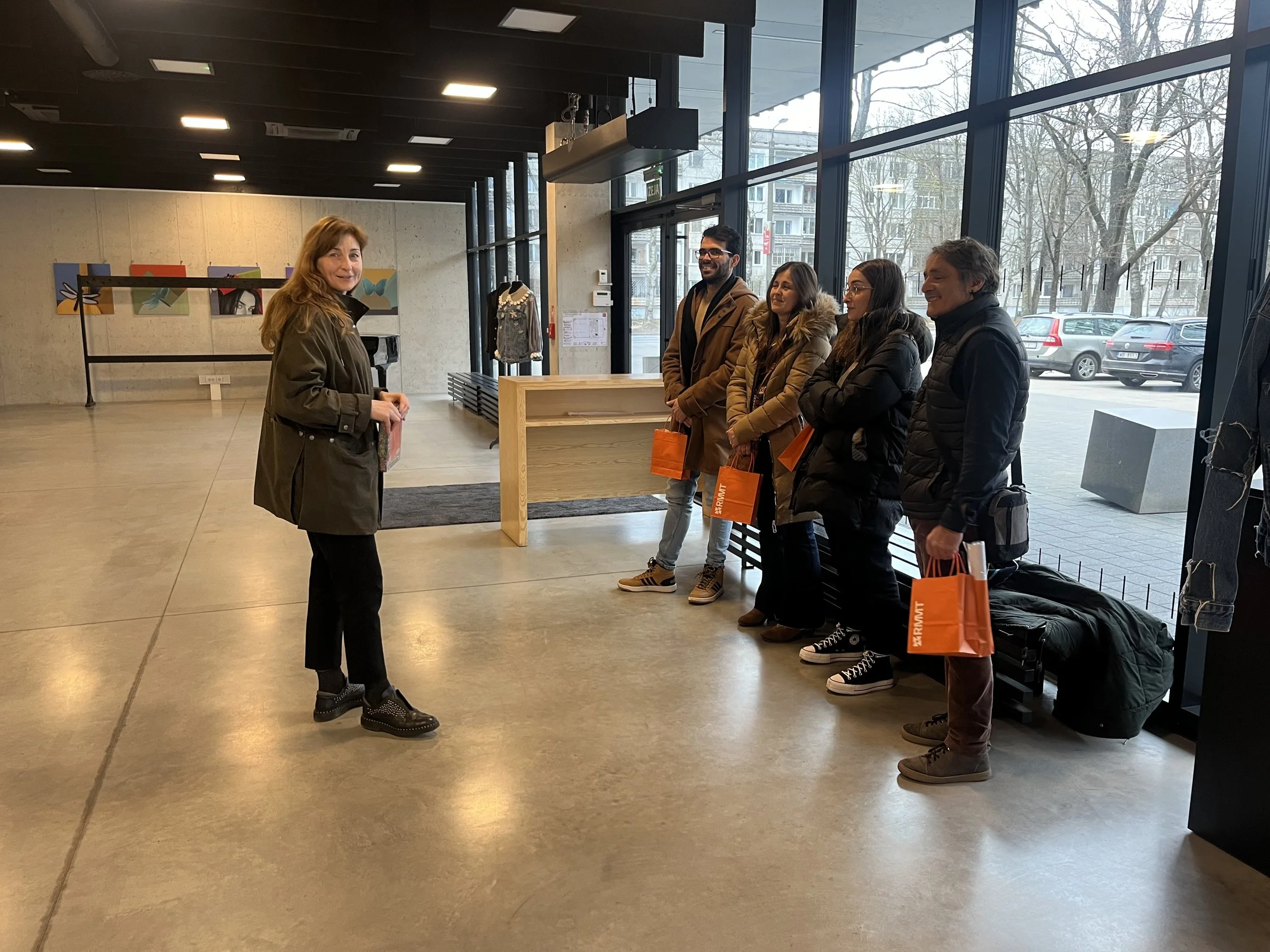 A woman standing and smiling in front of four people who are standing in a line inside a modern building with large glass windows, holding orange gift bags.