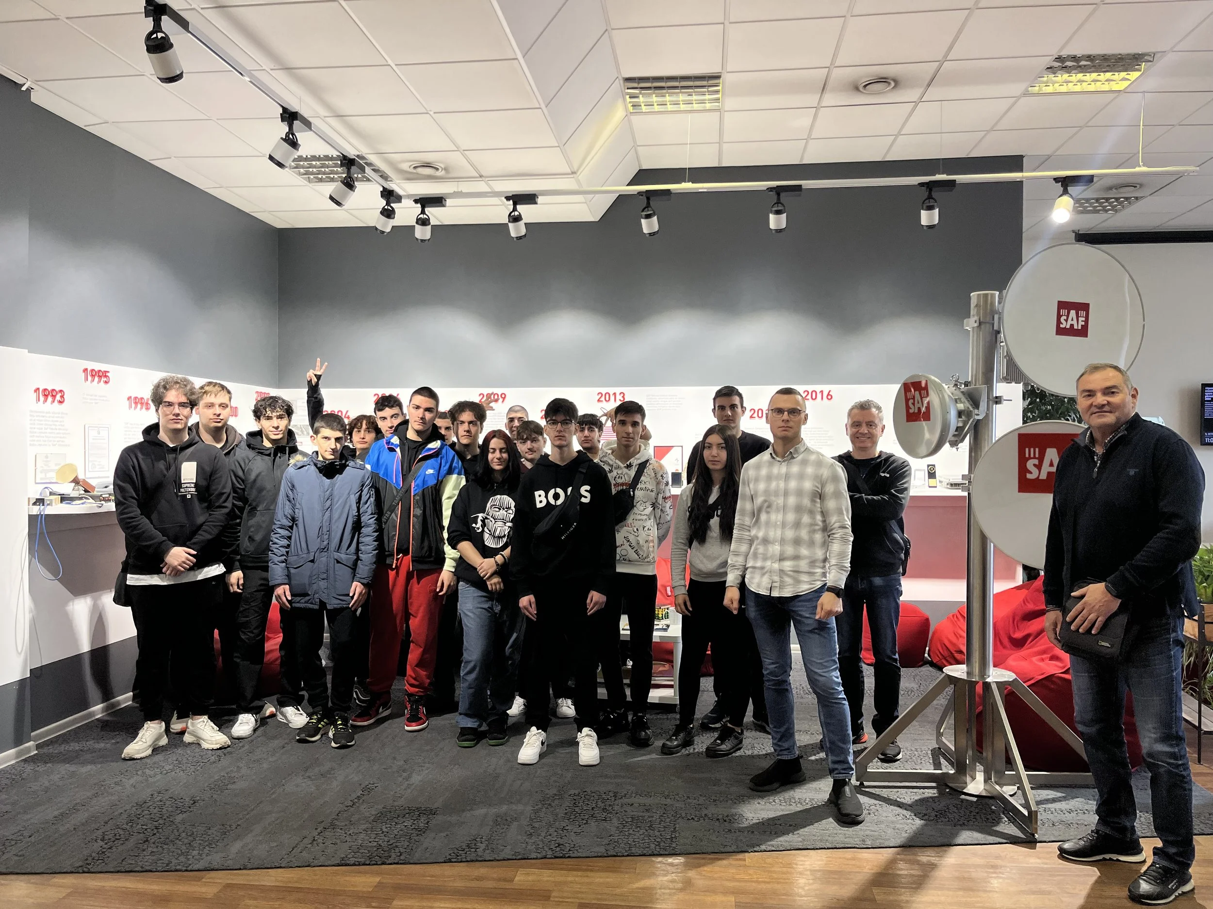 Group of young people and adults posing inside a museum or exhibition space, with display panels and large satellite dish antennas in the background.