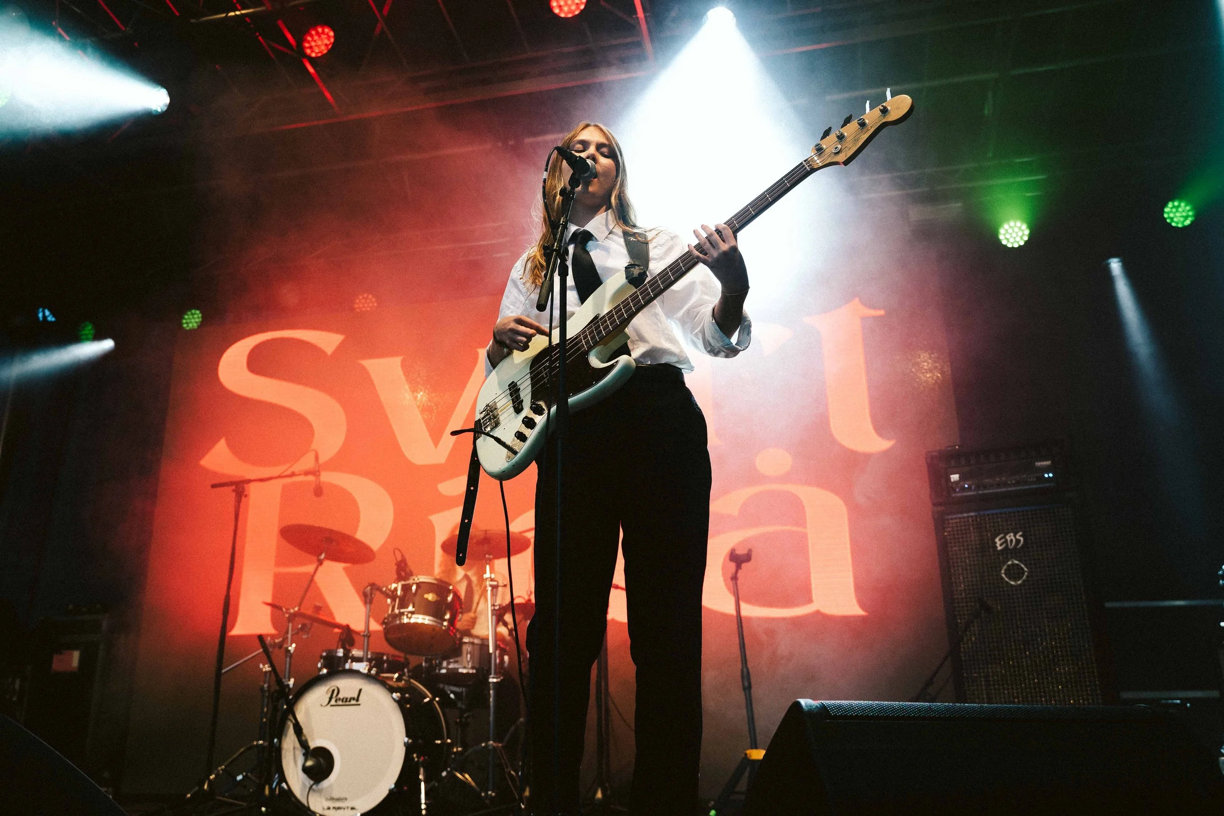 A woman playing bass guitar and singing into a microphone on stage, with a drum set and amplifier behind her, performing at a concert with colorful lighting and large text in the background.