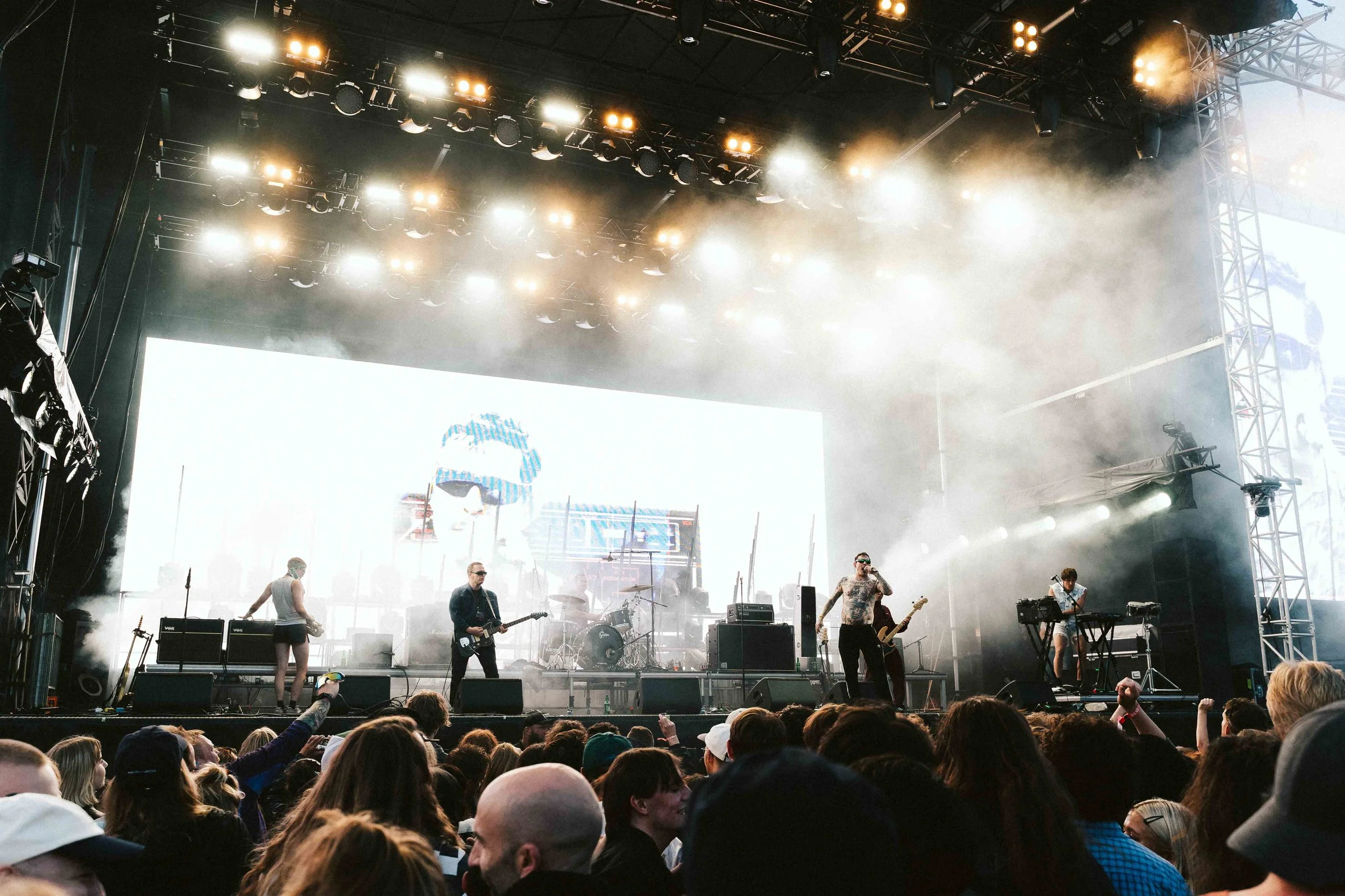 Band performing on an outdoor stage with smoke and bright lights, large LED screen in the background, and a crowd watching.