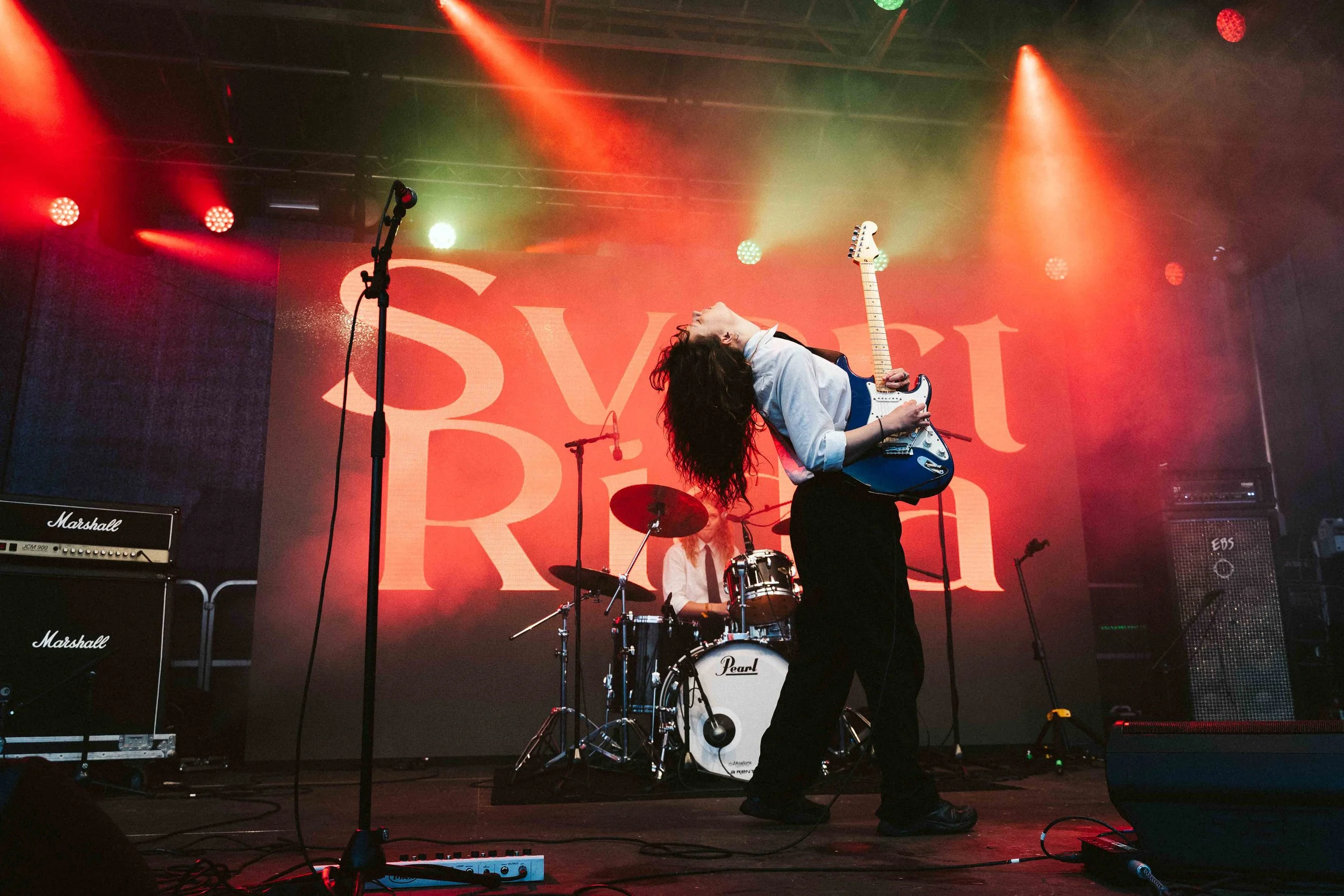 Musician playing electric guitar on stage with drummer in background during concert with red backdrop and colorful stage lights.