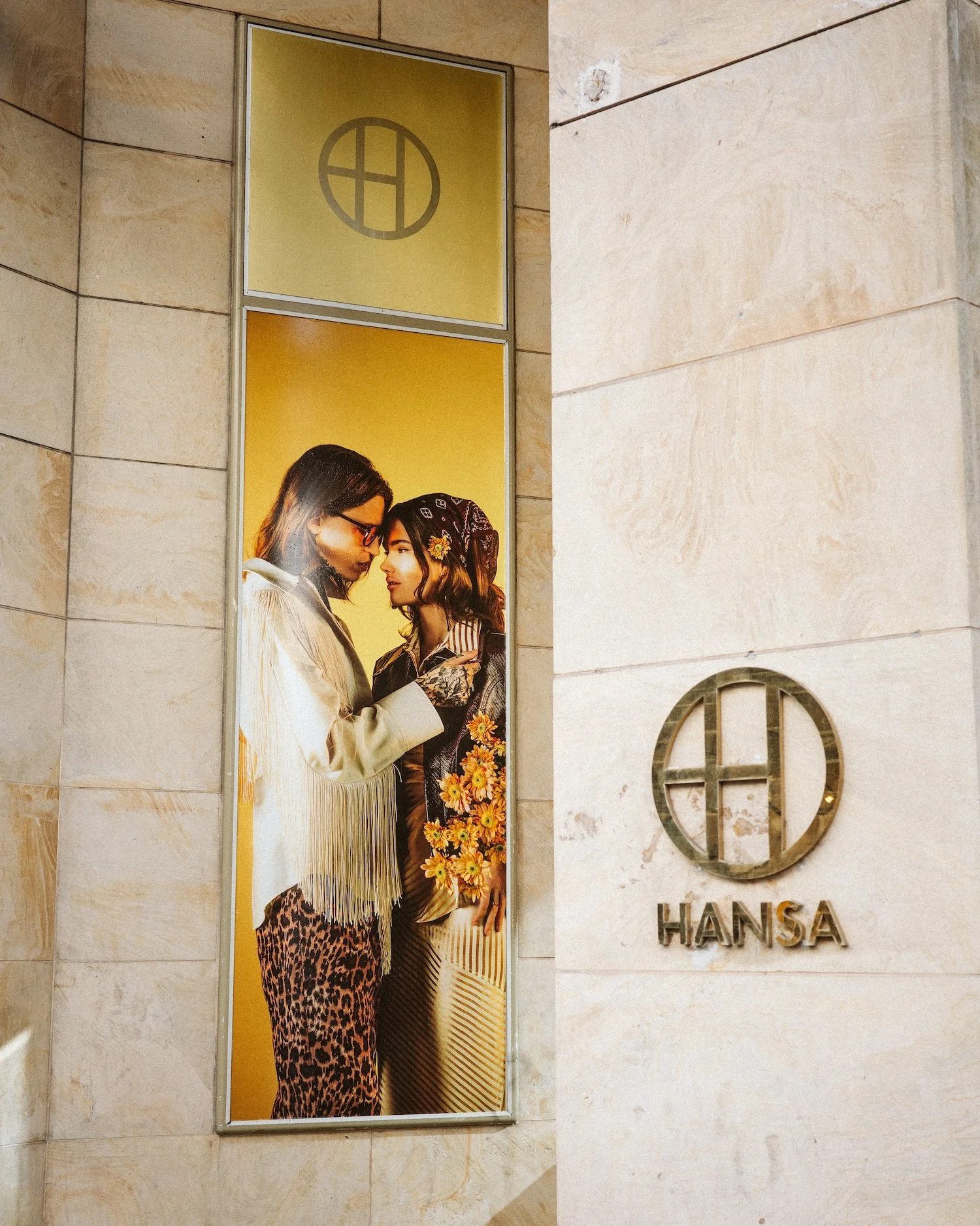 A fashion advertisement showing two women standing close, dressed in eclectic, stylish clothing with floral and animal print details, inside a shopping mall or store with the sign 'HANSA' on the wall.