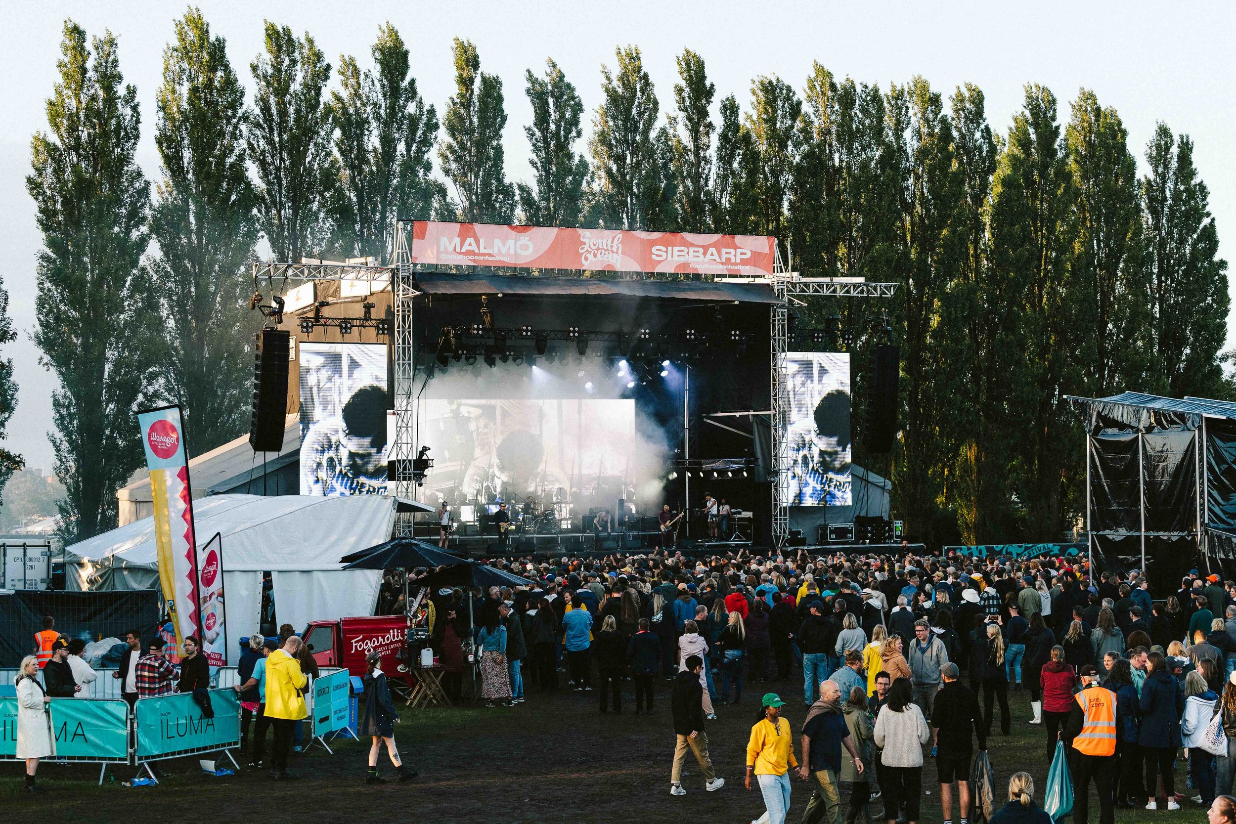 Large outdoor concert stage with band performing, big screens displaying performer, crowd gathered watching, trees in background, some people walking, event banners, and security personnel.