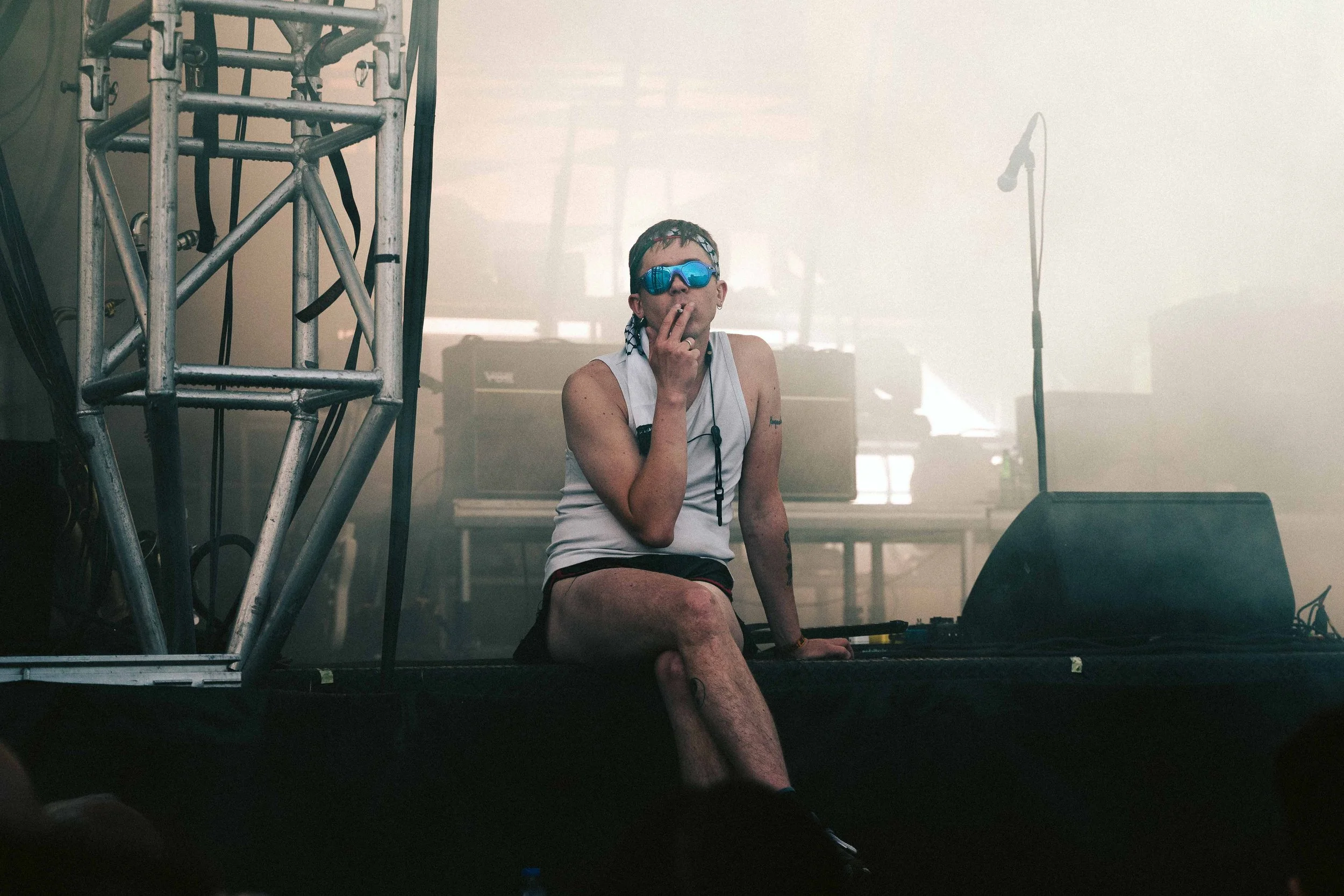 A person with short hair, wearing sunglasses and a sleeveless shirt, sitting on stage near sound equipment, with a microphone stand nearby, in a smoky indoor setting.