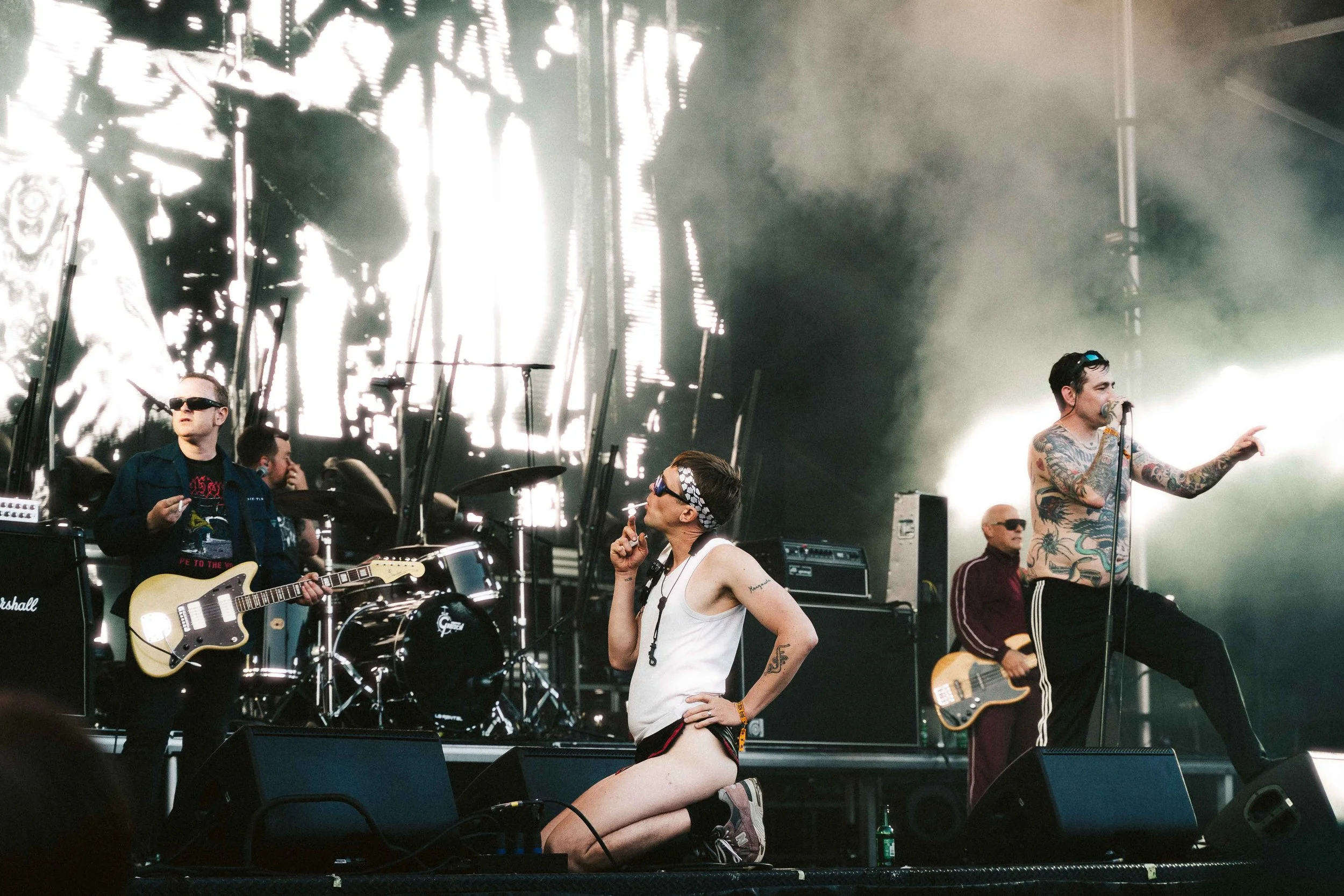 A band performing on stage with a singer kneeling in front, a guitarist on the left, a bassist on the right, and a drummer in the background. The singer has tattoos and is gesturing with his left hand. The stage has bright lights and a large screen b