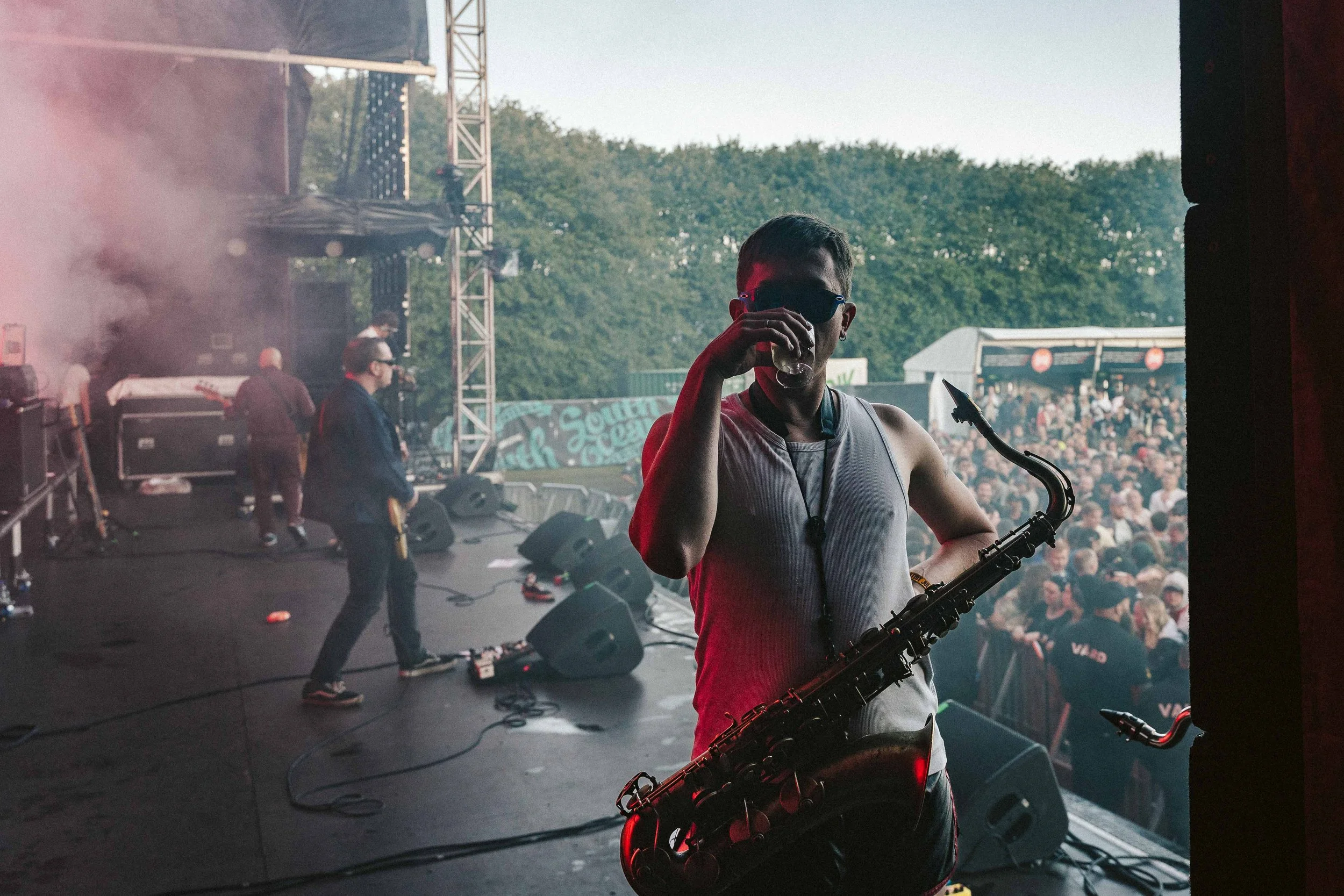 A musician with sunglasses and a sleeveless shirt drinking water on stage at a concert, holding a saxophone, with a crowd and stage setup in the background.