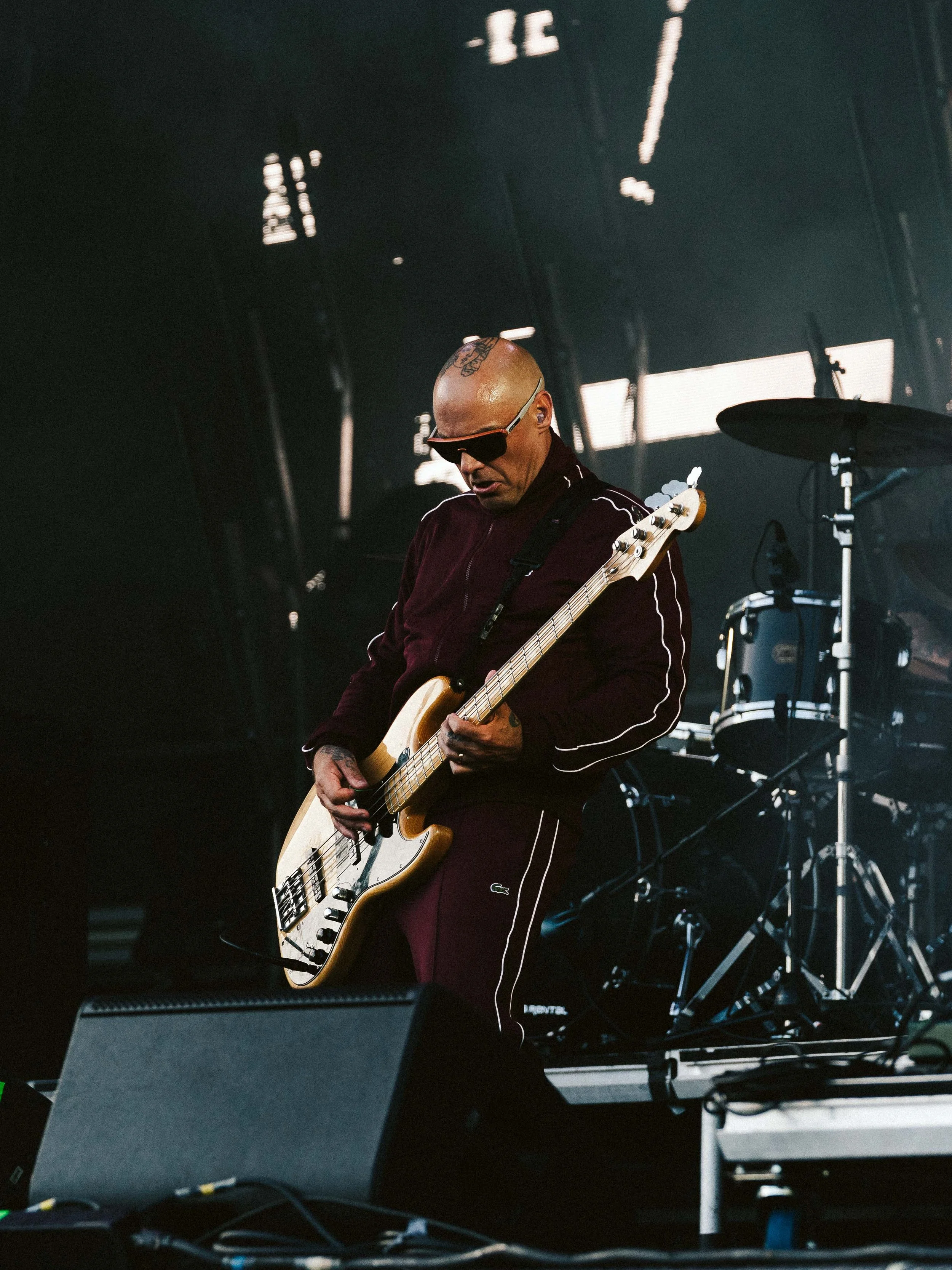 Musician playing electric bass guitar on stage at a concert, wearing sunglasses and a dark tracksuit, with drums in the background.