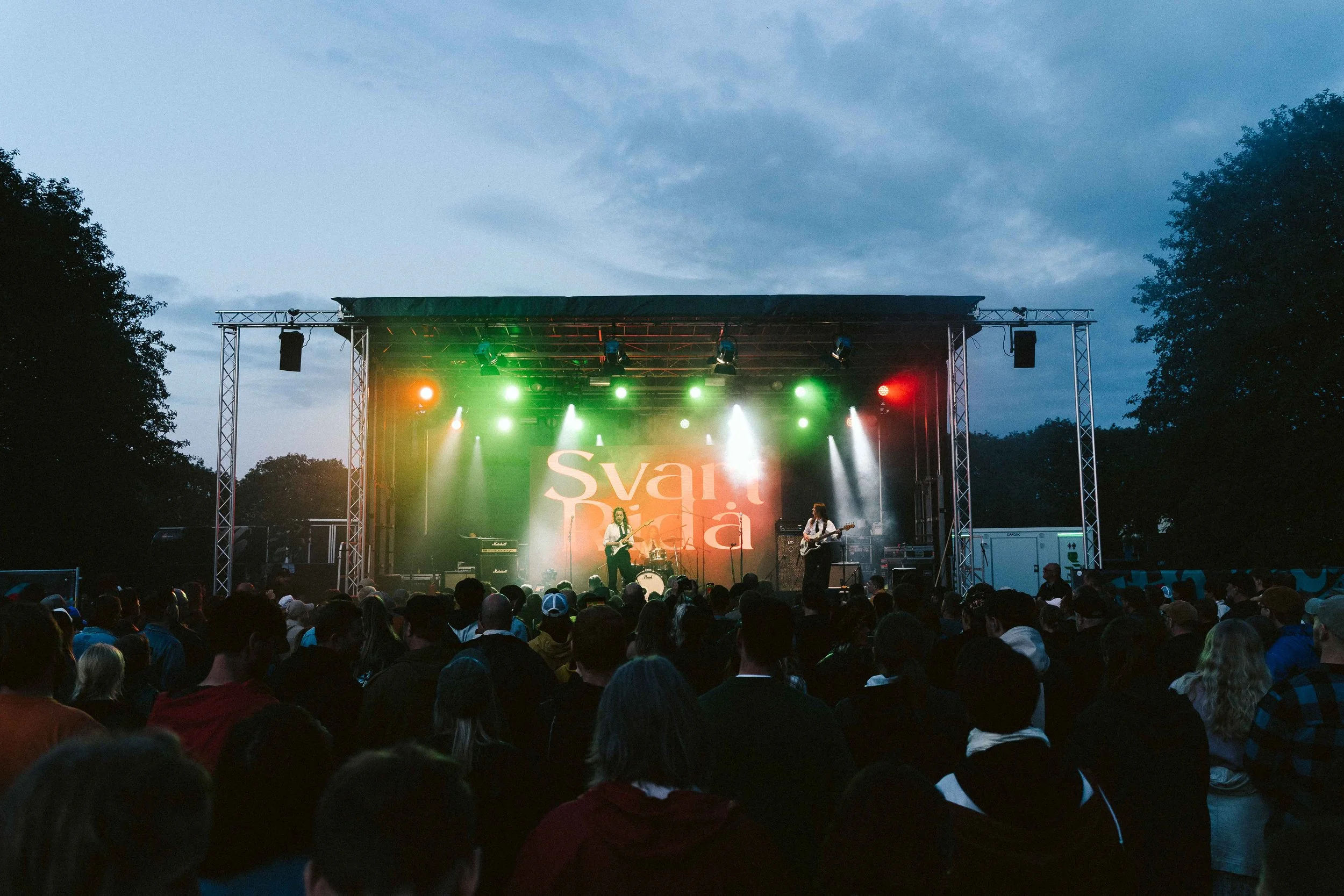 An outdoor concert at dusk with a band performing on stage under colorful lights, and a large crowd watching.