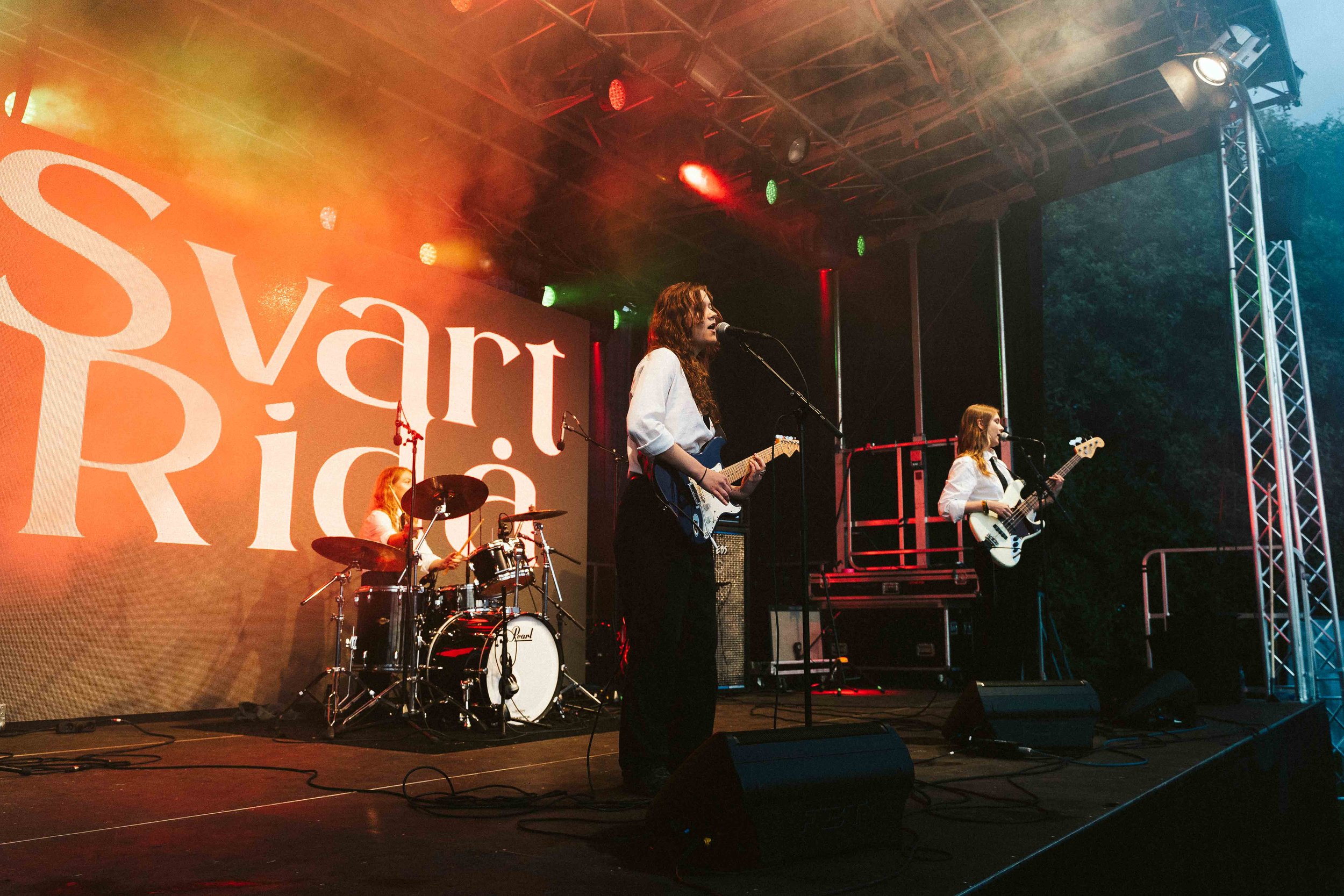 A three-member band performing on stage at a concert or festival during evening. The drummer, guitarist, and bassist are all women, dressed in white shirts. The stage has orange, green, and red lighting with a large screen behind displaying partial w
