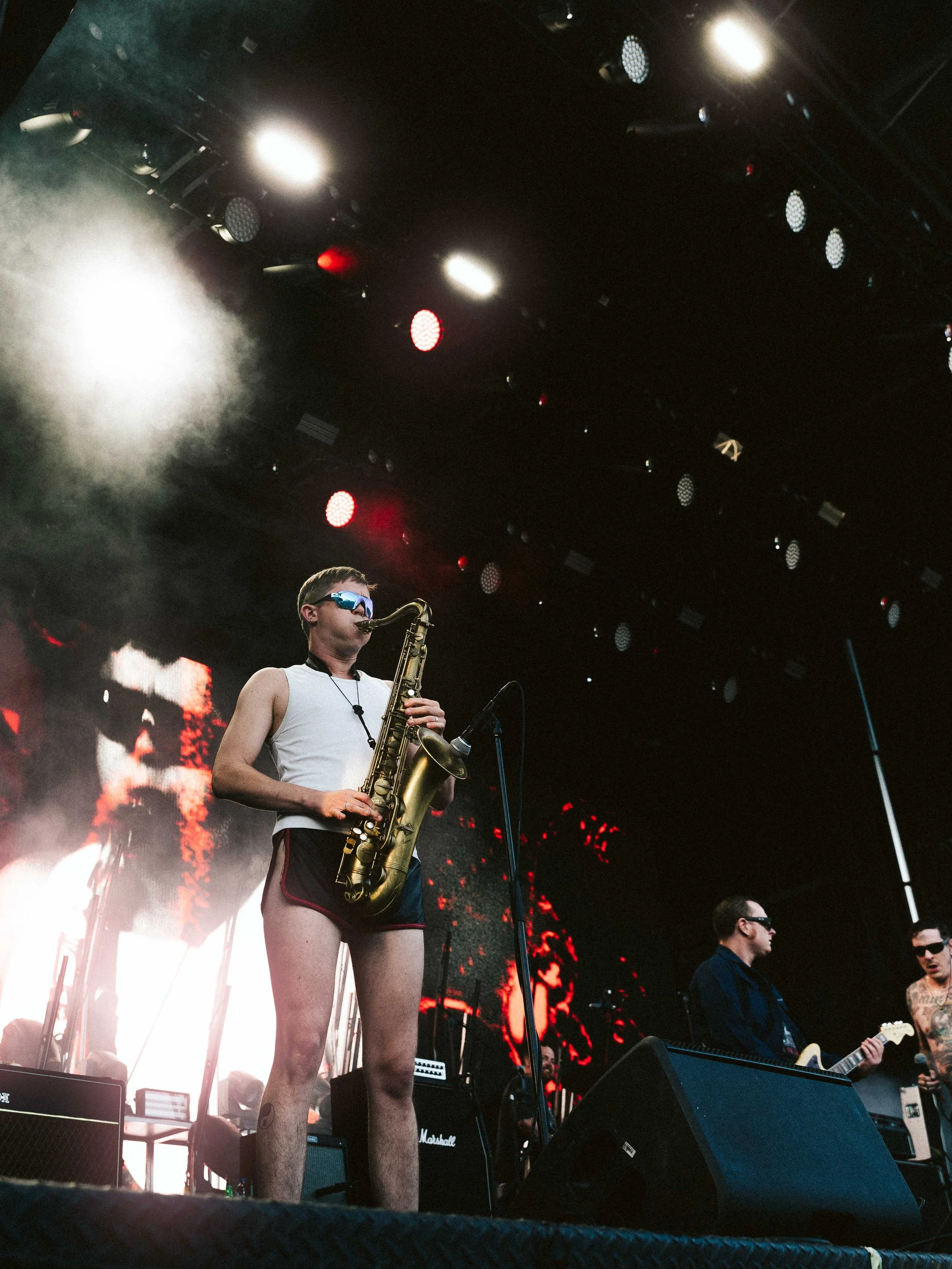 Young musician wearing sunglasses and shorts playing a saxophone on stage with band members and large screen in background.