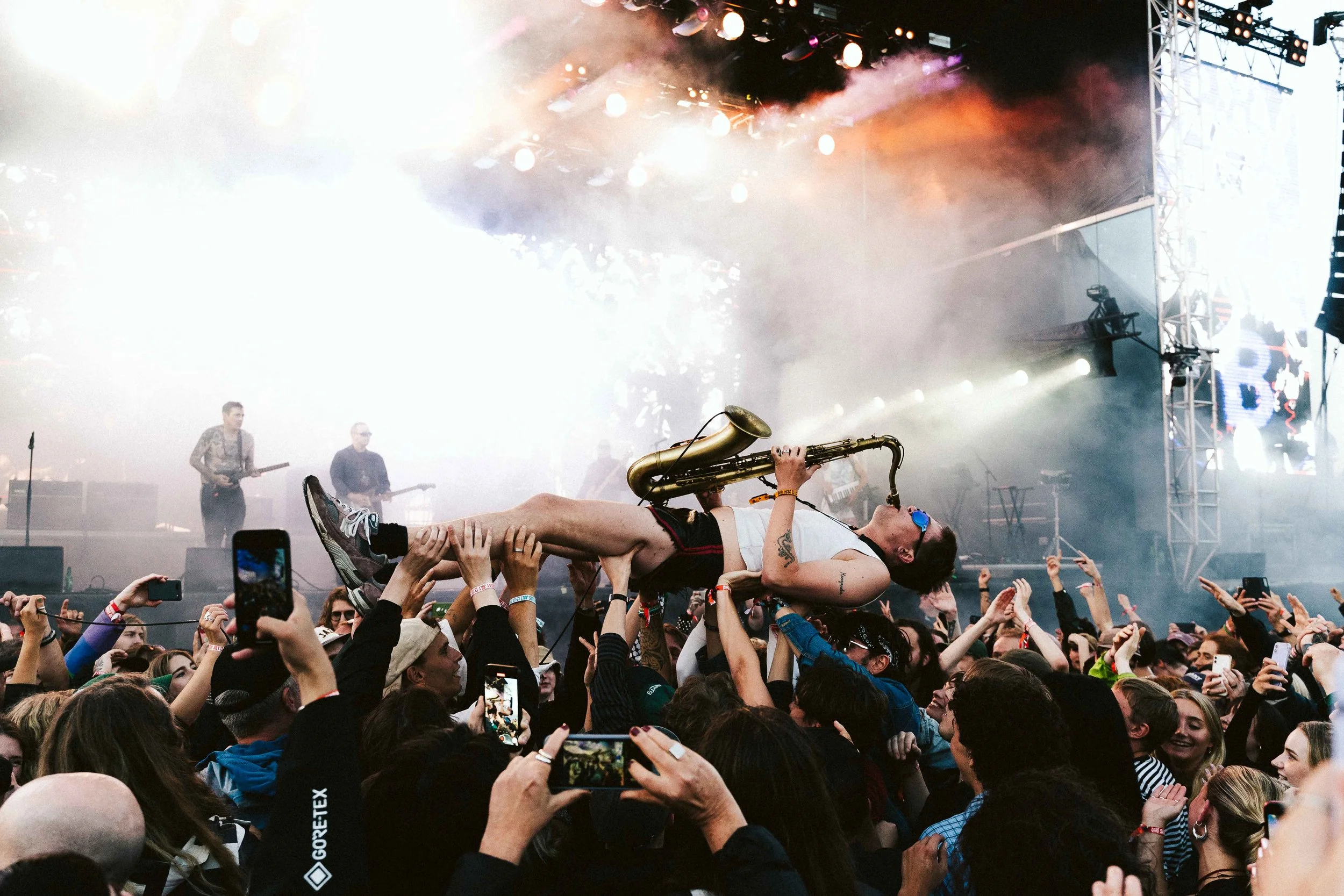 A musician with sunglasses and tattoos is lying across the crowd at a concert, playing a saxophone while crowd-surfing. The stage in the background is lit with bright lights and fog, with other band members playing instruments.