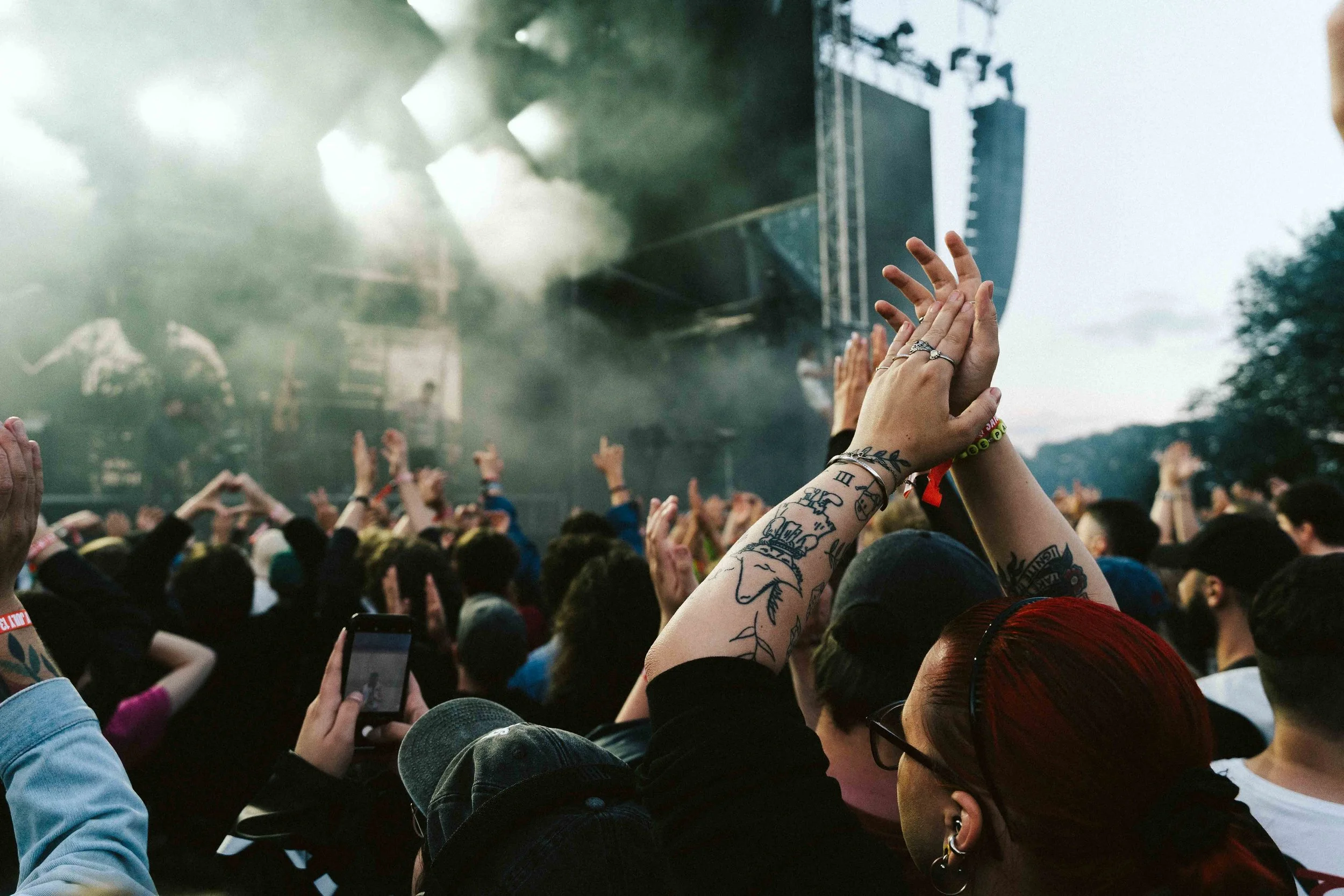 Crowd at a concert or festival with hands raised, some clapping or taking photos, on an outdoor stage with smoke and lighting effects.