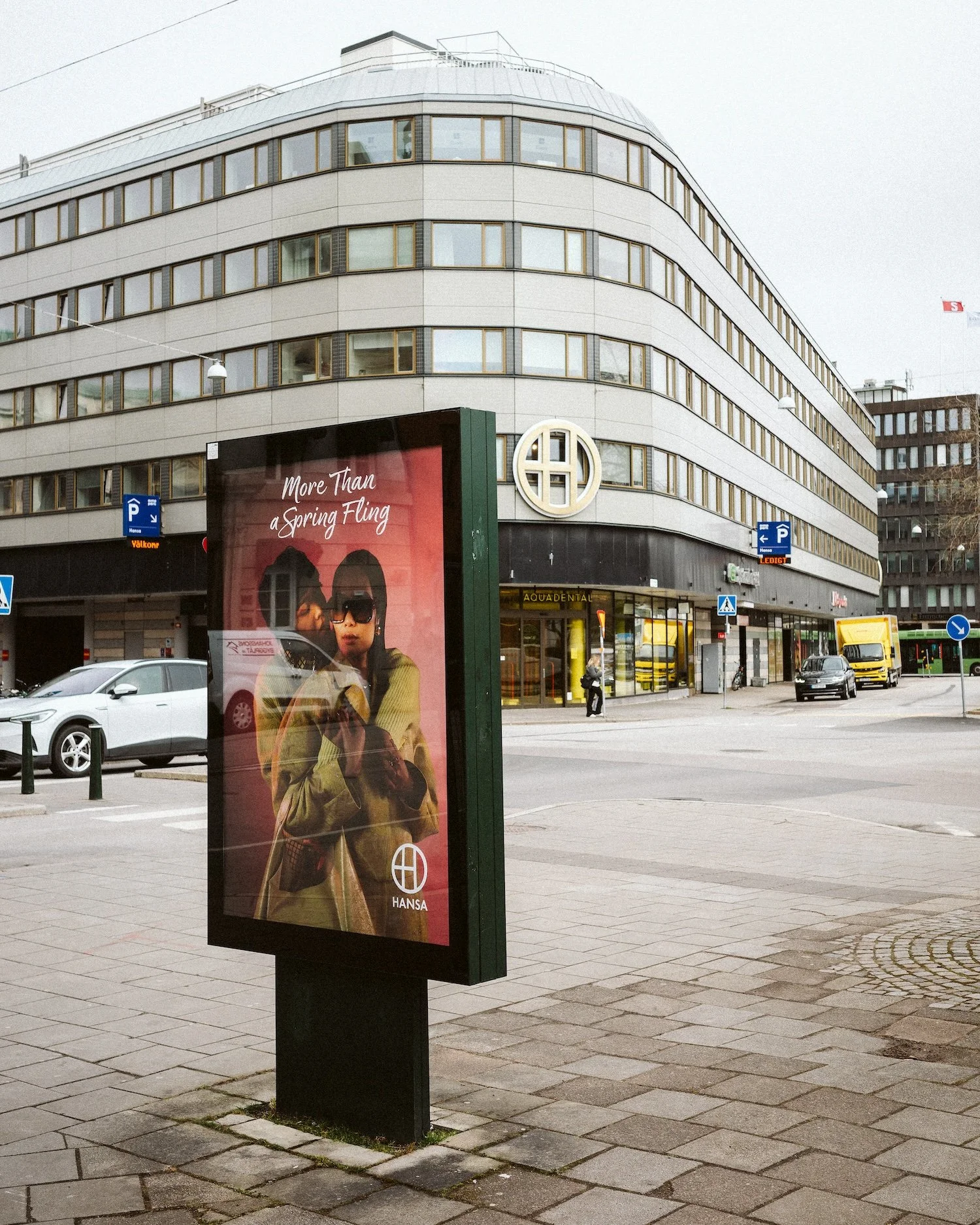 City street scene with a large building, parked cars, a yellow delivery truck, and a digital billboard featuring an advertisement with a woman wearing sunglasses and a coat, with the text 'More Than a Spring Fling'.