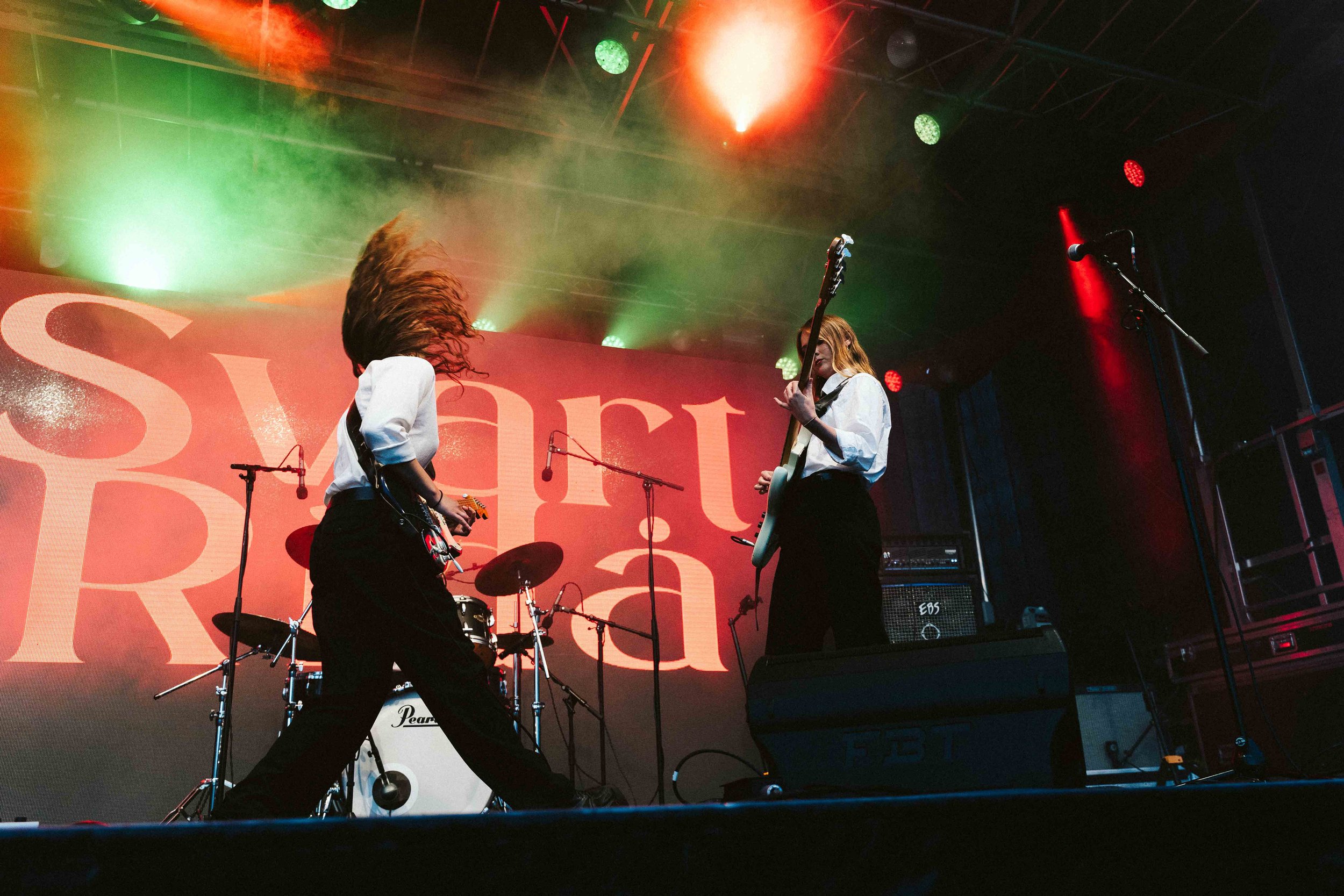 Two female musicians performing on stage with guitars and drums, with colorful stage lighting and a large red backdrop displaying the words 'Swarat'.