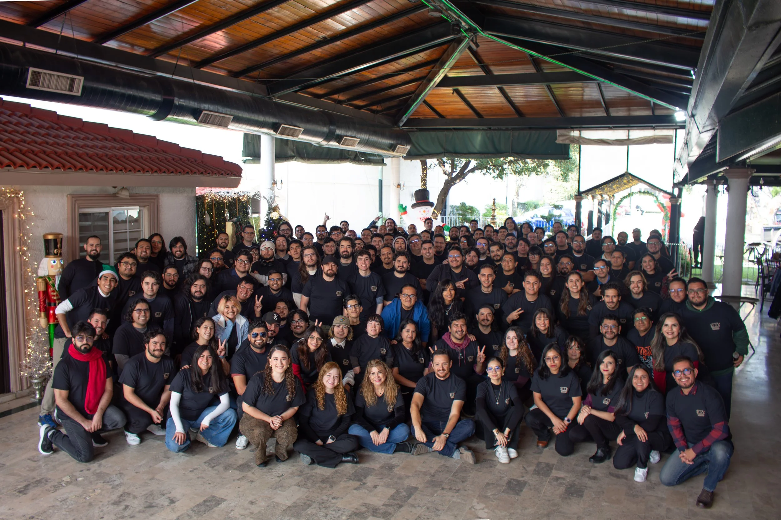 A large group of people posing for a photo indoors decorated with Christmas ornaments, including a snowman figure, Christmas trees, and string lights.