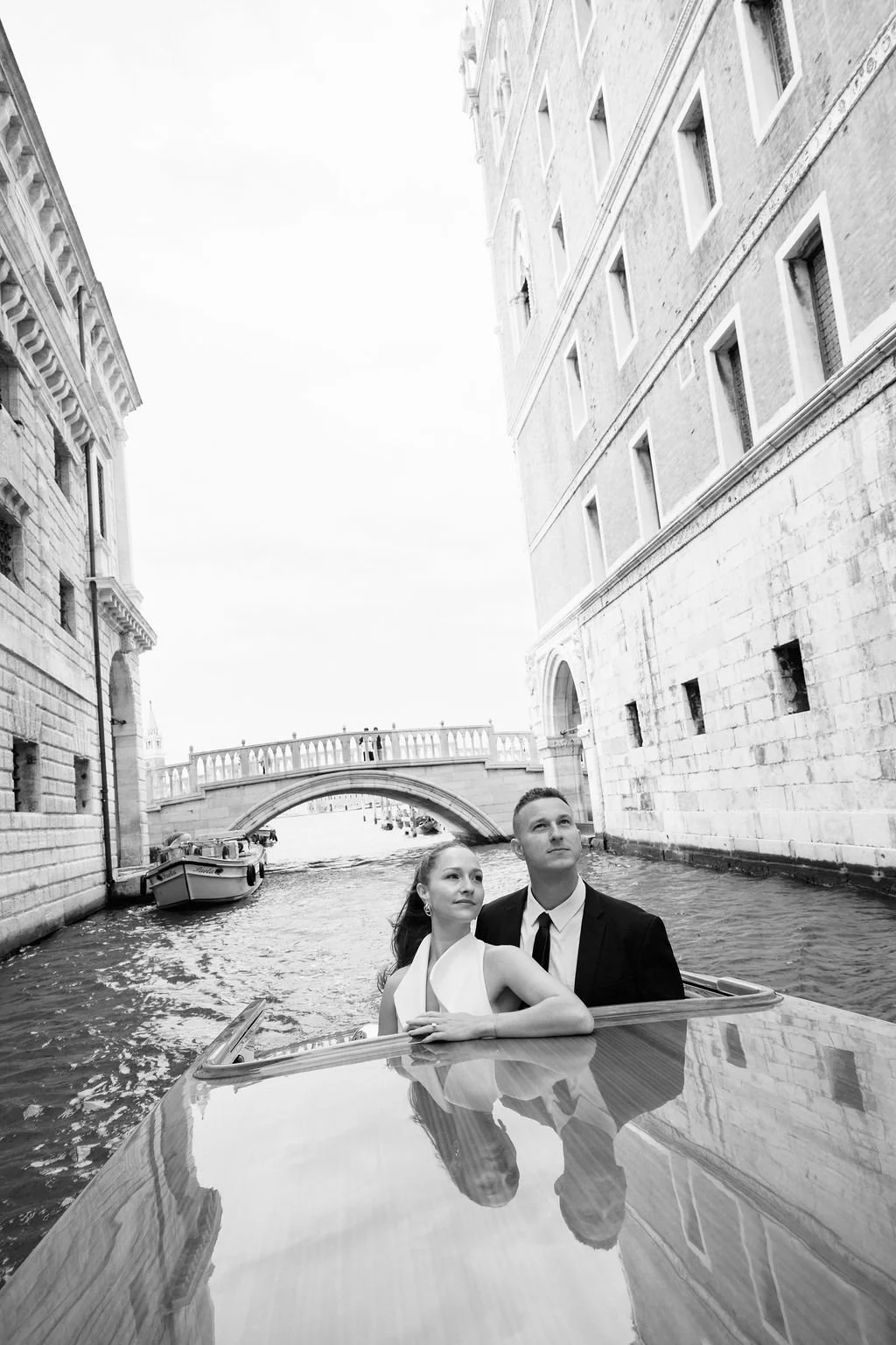 A black-and-white photo of a couple on a boat ride through a canal in Venice, Italy, with historic buildings on either side and a bridge in the background.