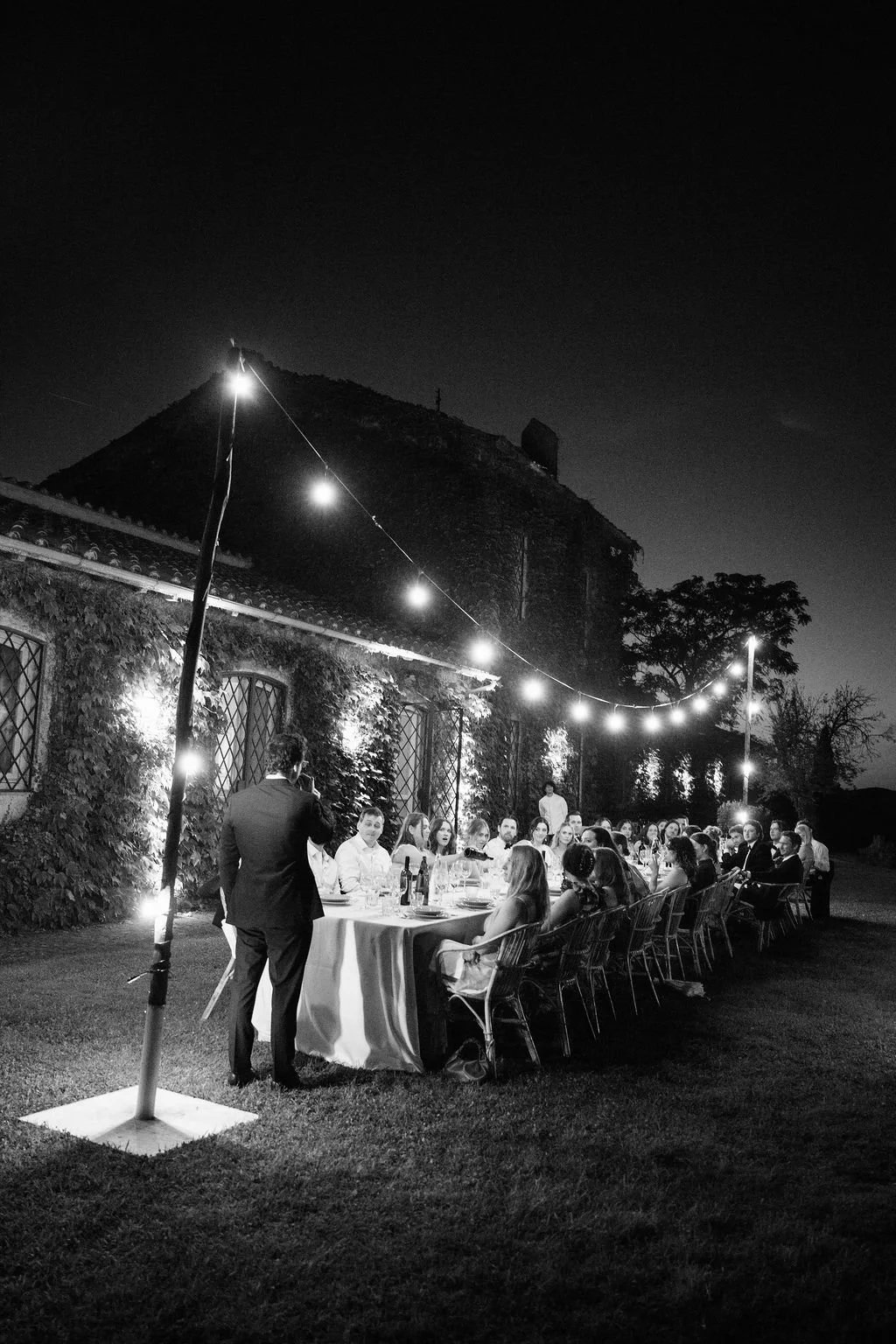 Outdoor dinner party at night with long table of guests under string lights, historic building and trees in background, in black and white
