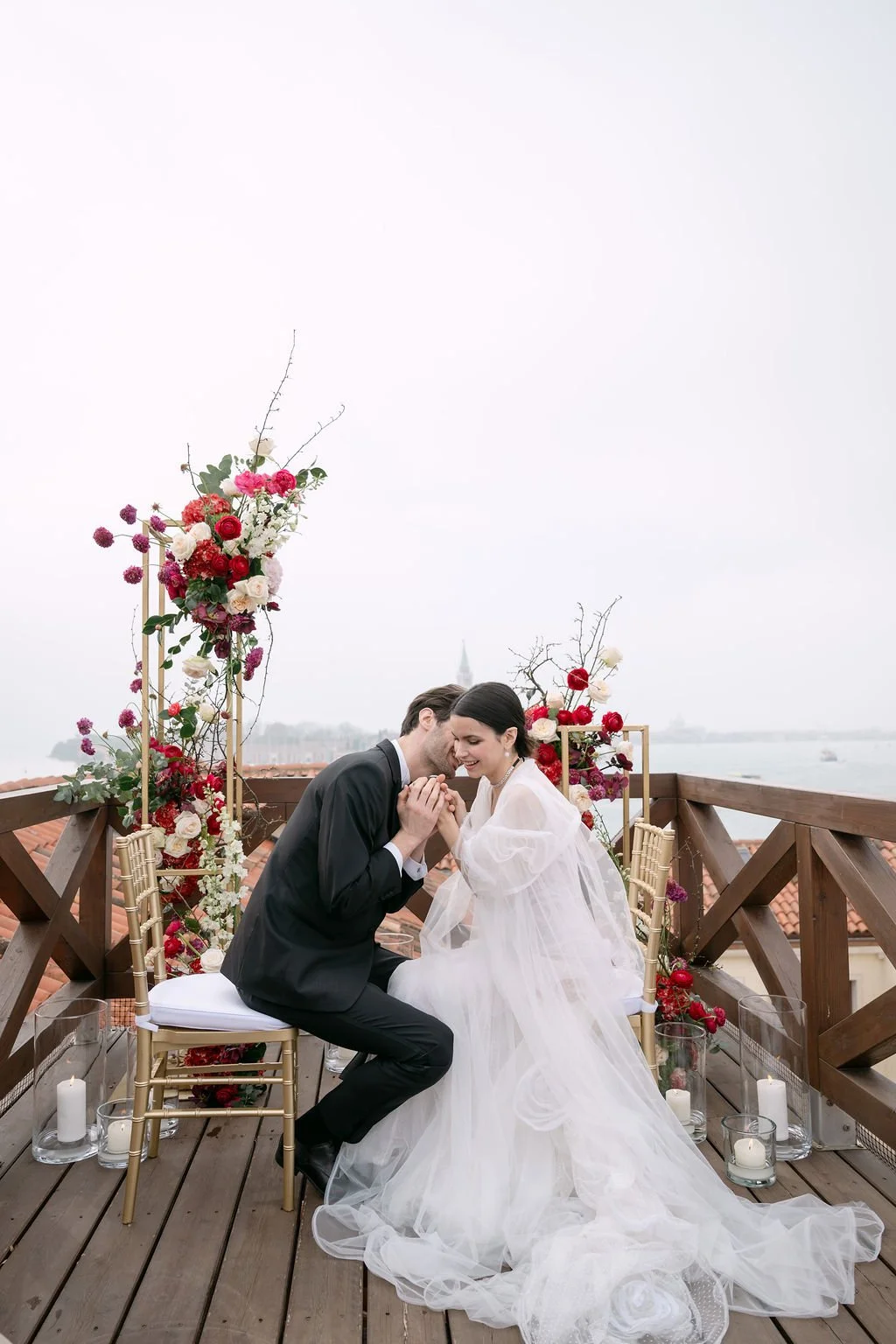 A couple dressed in wedding attire on a decorated outdoor balcony, sharing a tender moment.