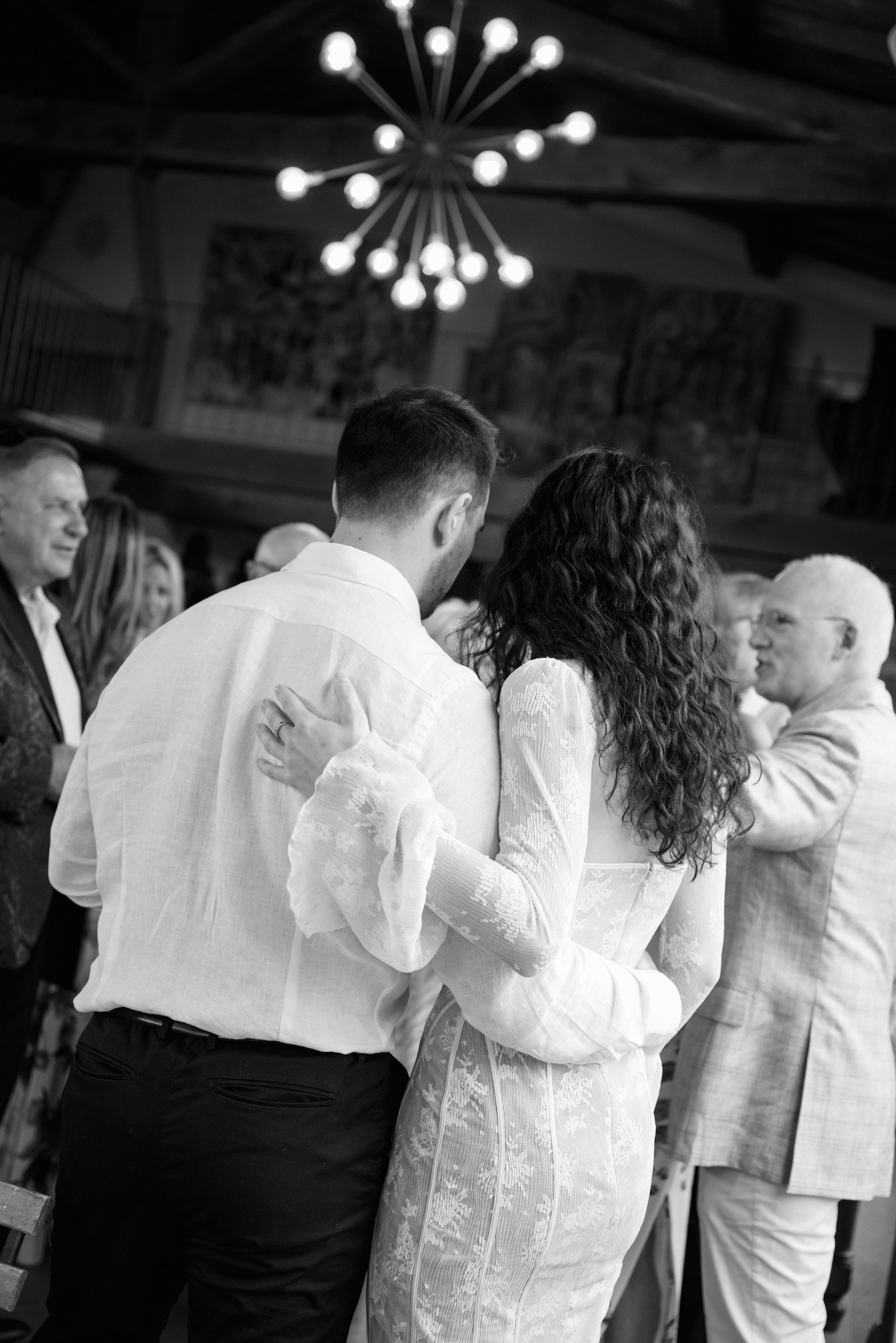 A black and white photo of a couple dancing at a celebration, surrounded by guests, with a modern chandelier hanging from the ceiling.