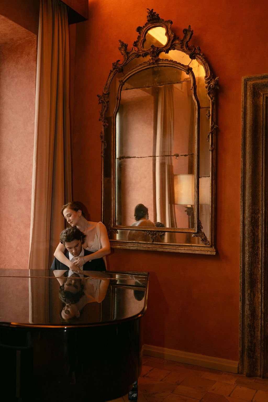 A woman embraces a man at a grand piano in a warmly lit room with a large ornate mirror and reddish walls.