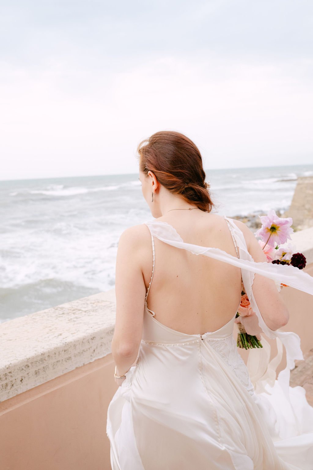 A woman in a white dress holding flowers by the seaside, with her back turned to the camera and ocean waves in the background.