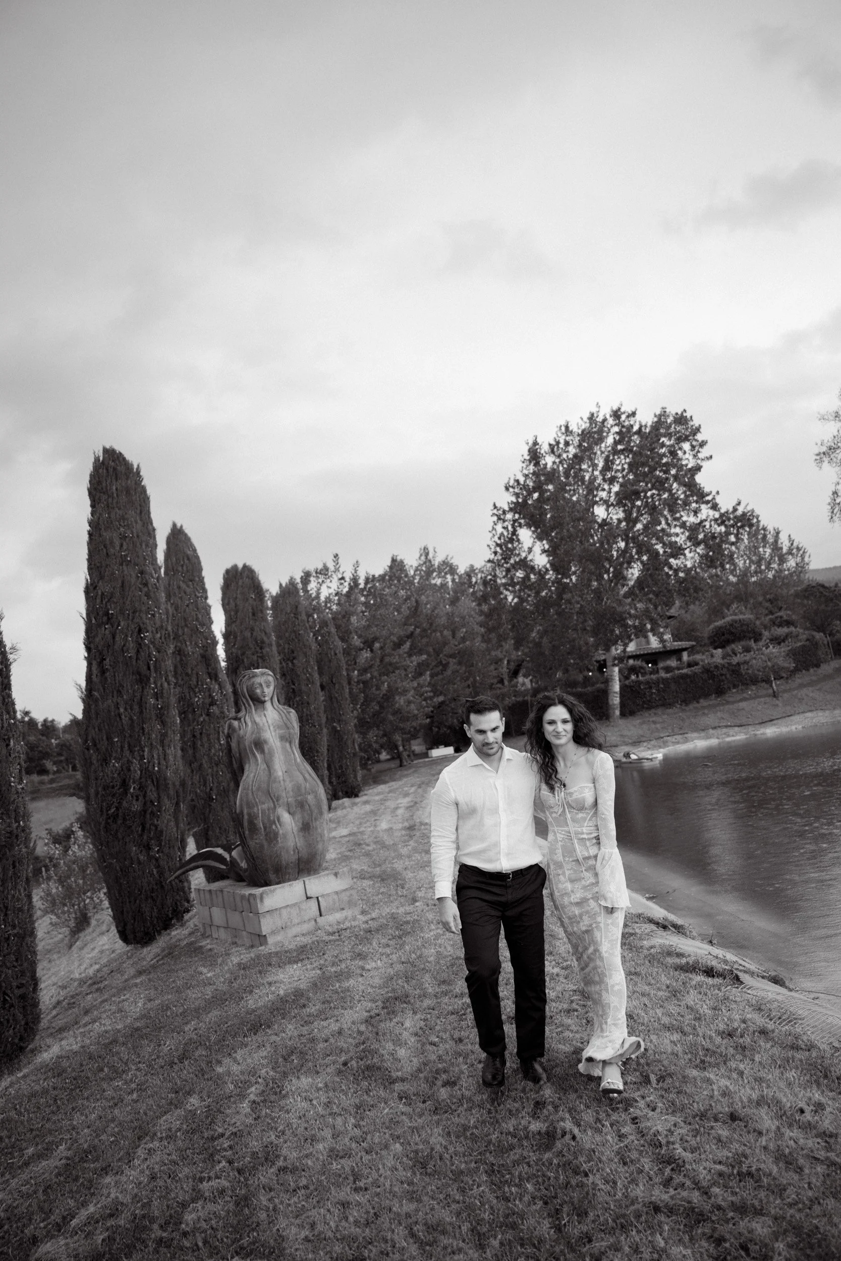 A black and white photo of a man and woman walking along a grassy path beside a lake. The woman wears a long lace dress, and the man wears a white shirt and dark pants. Tall trees and a sculpture of a woman are visible in the background.