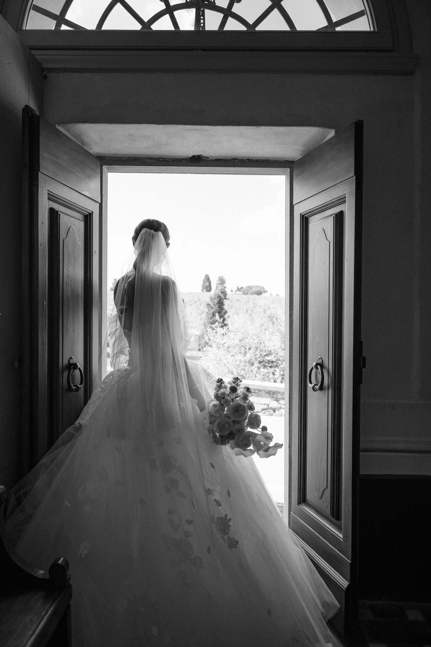A bride in a wedding gown and veil holding a bouquet of flowers, standing in the doorway of a church, looking out to a scenic outdoor landscape.