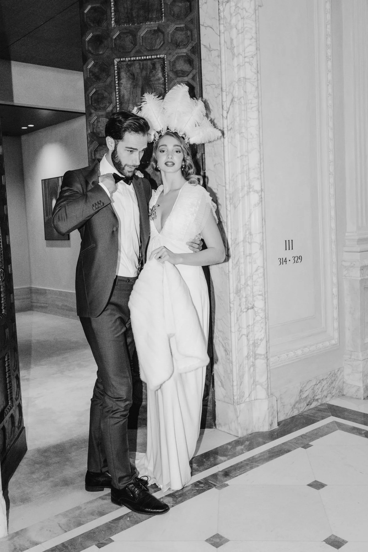 A black and white photo of a man in a tuxedo and a woman in a white dress with feathered headwear, standing inside a building with marble walls.