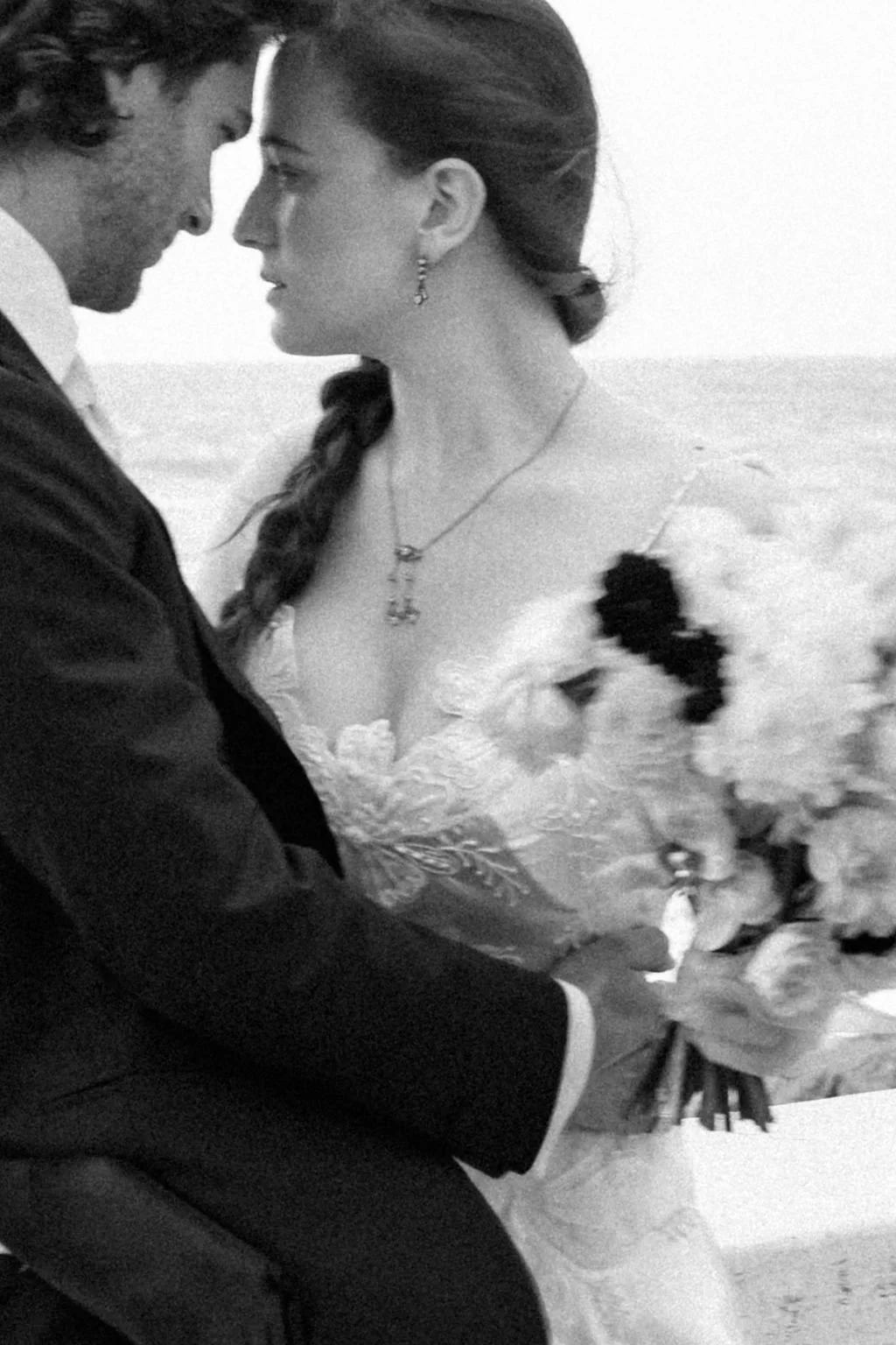 A black and white photo of a couple in wedding attire, with their foreheads touching and eyes closed, holding a bouquet of flowers, outdoors.