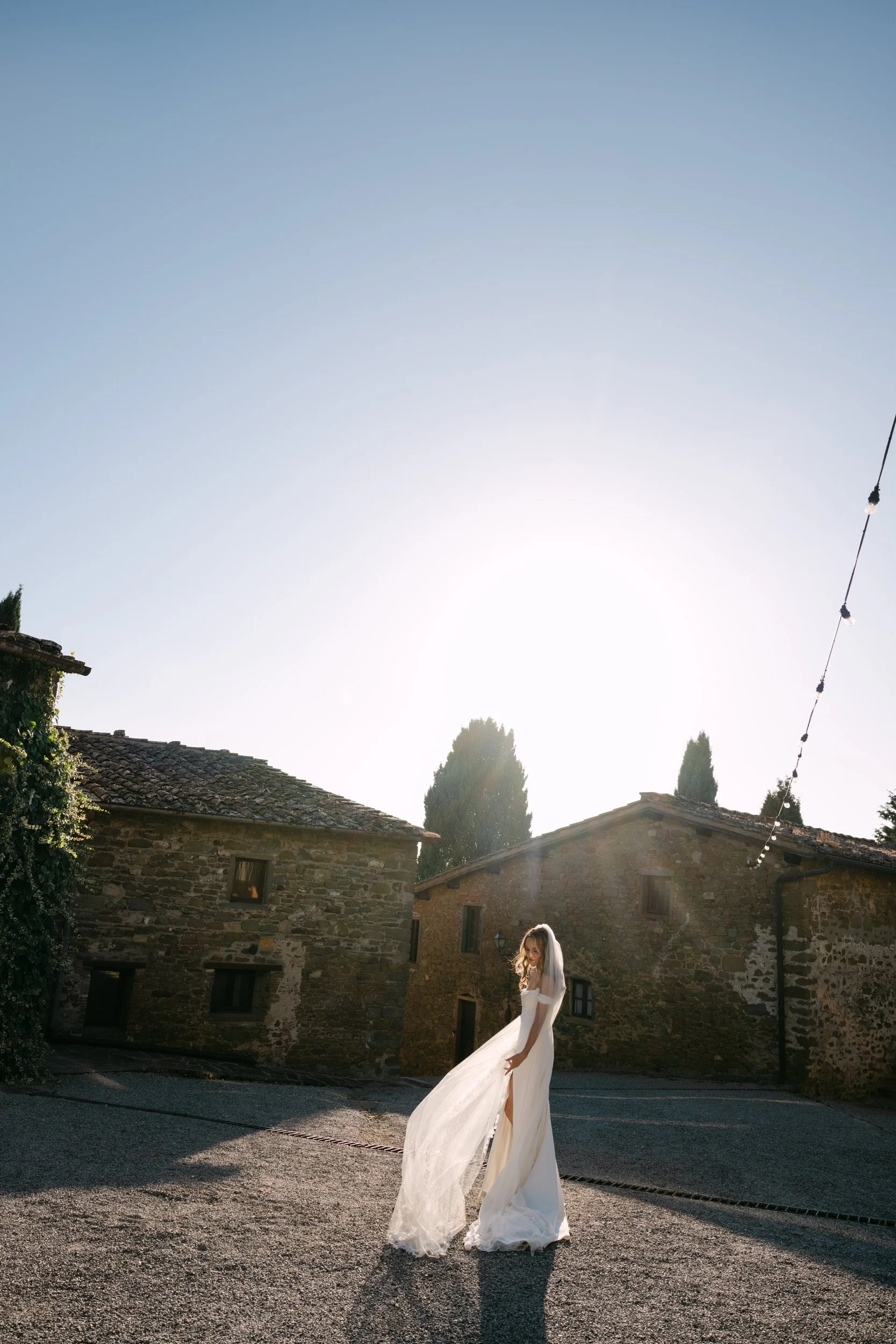 A bride in a white wedding gown with a veil standing outdoors on a cobblestone street, with stone buildings and trees in the background, under a bright sky.