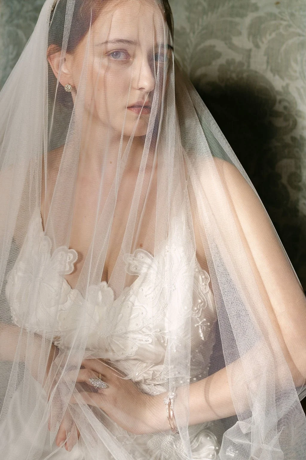A woman wearing a wedding dress and veil, with pearl earrings and a ring, looking away behind a sheer veil.