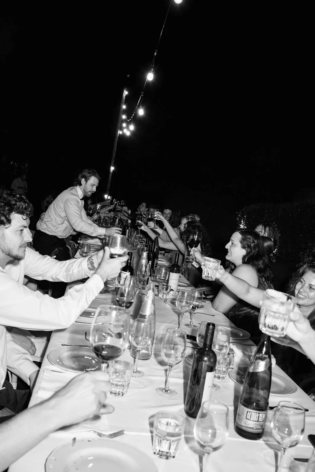 People sitting at a long outdoor dinner table raising glasses in a toast, under string lights at night.