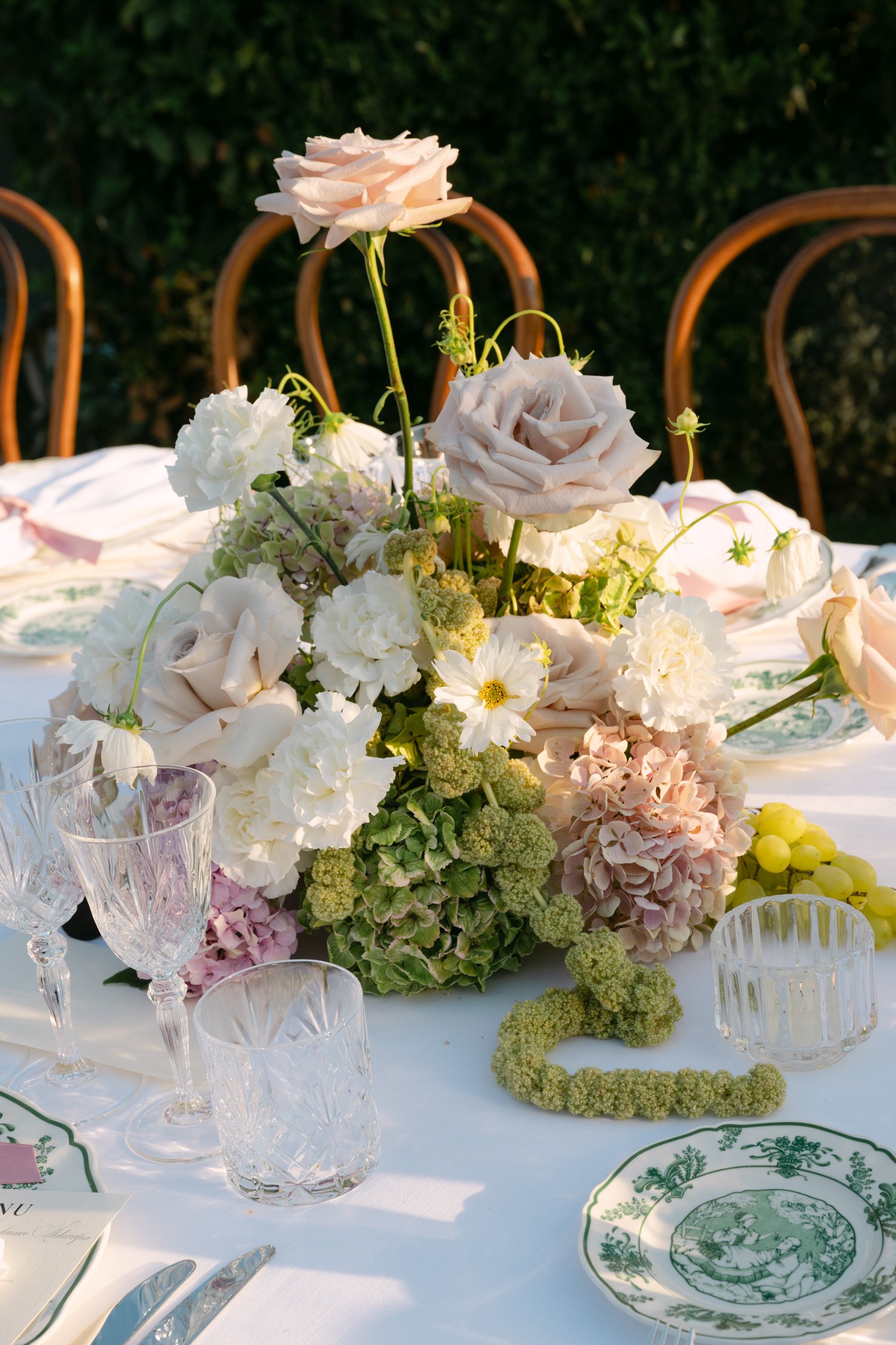 A floral centerpiece with large pink and white roses, hydrangeas, and other flowers on a white tablecloth, set on an outdoor dining table surrounded by empty glasses and plates.