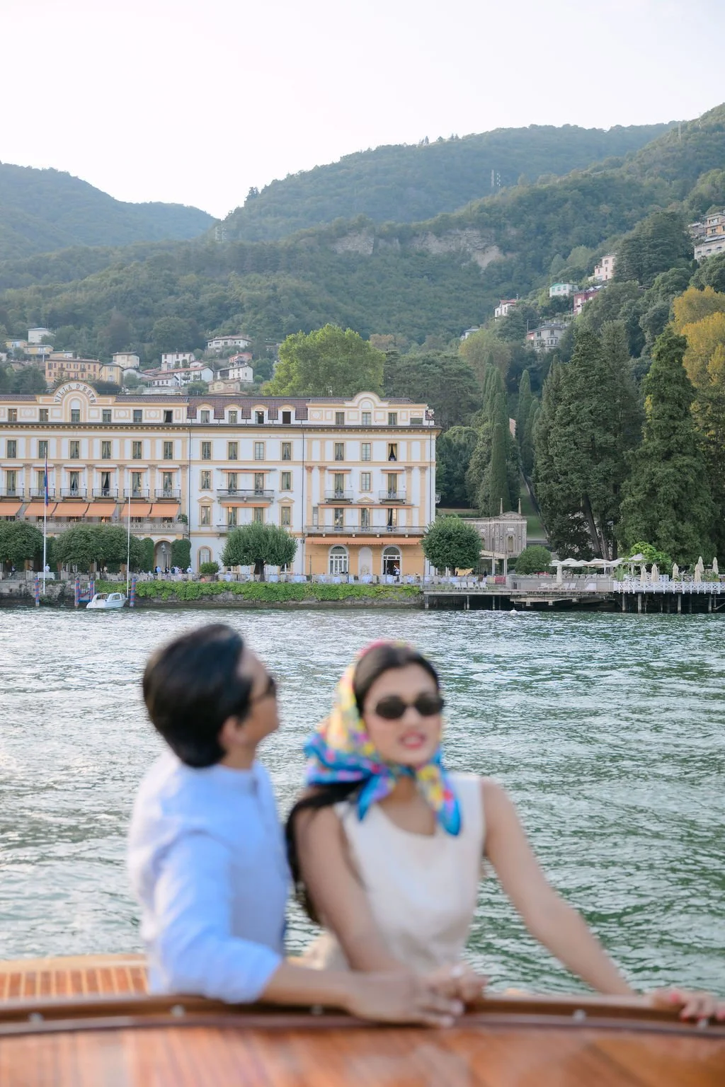 Two women sitting on a boat on a river, with a large ornate building and green hills in the background.