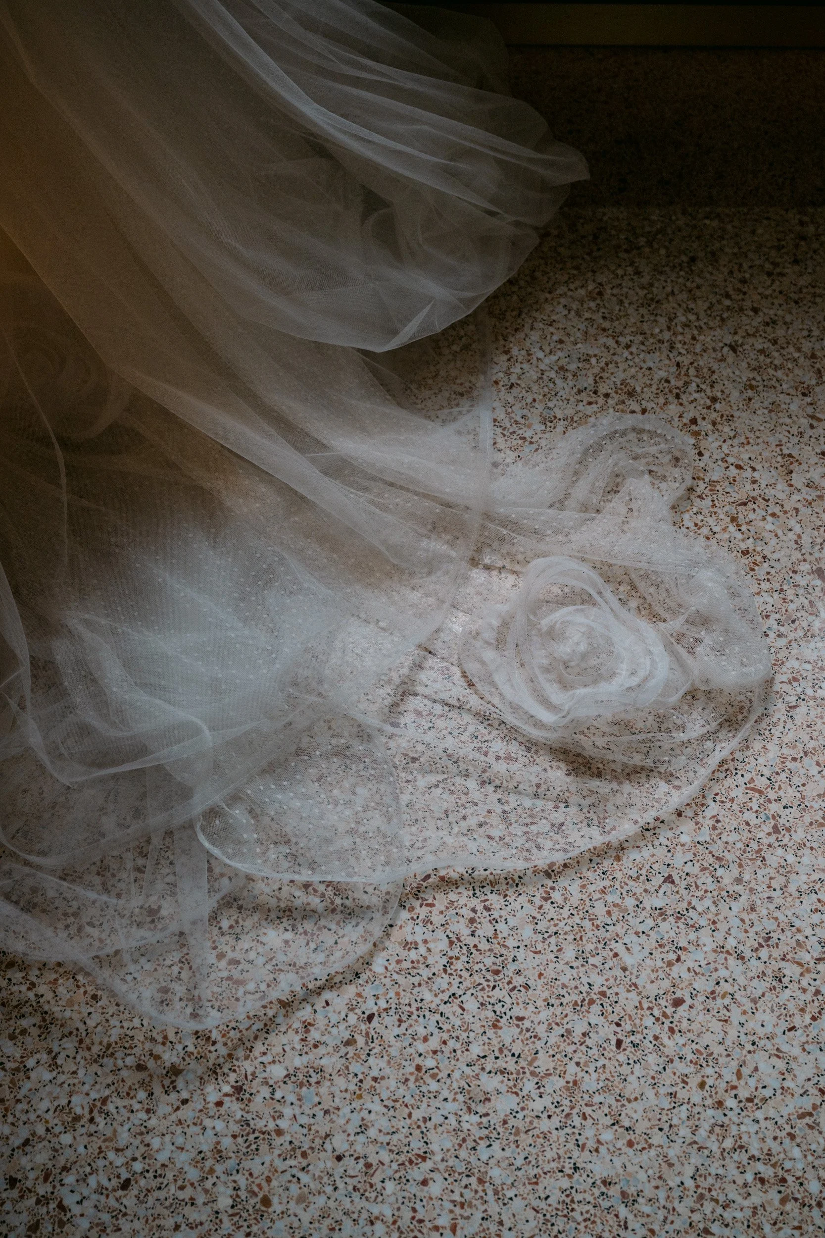 A white, sheer, polka-dotted veil with a floral applique on a speckled tile floor.
