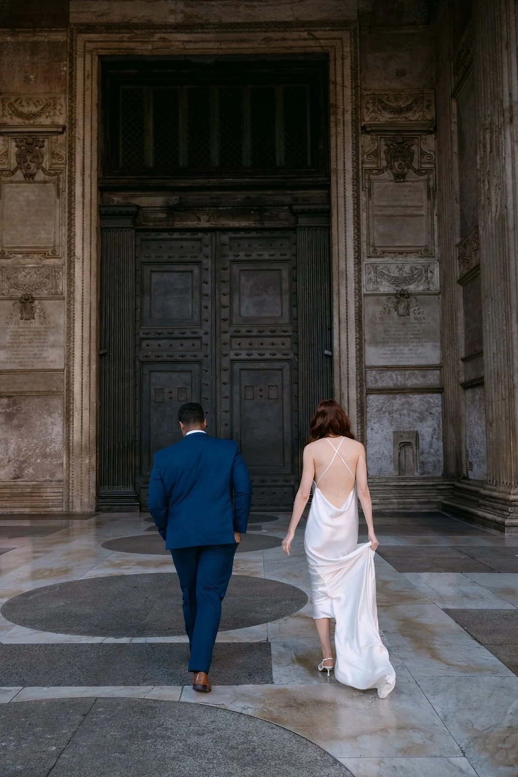 A man and a woman dressed in formal attire walking towards a large ornate door in a historic building with intricate stone carvings.