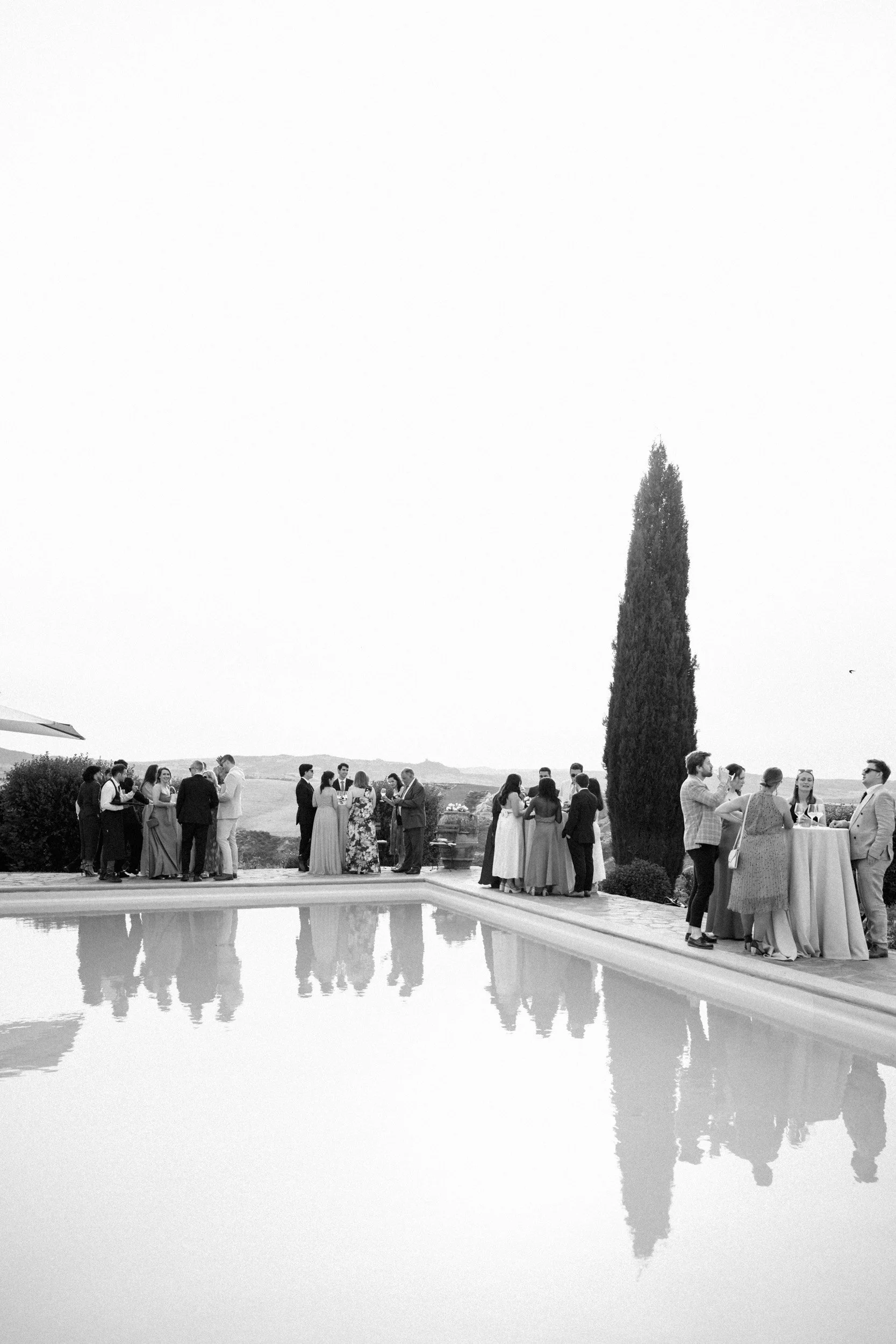 People gathered outdoors by a pool, socializing and taking photos, with a large cypress tree and a scenic landscape in the background, during what appears to be a celebration or event.
